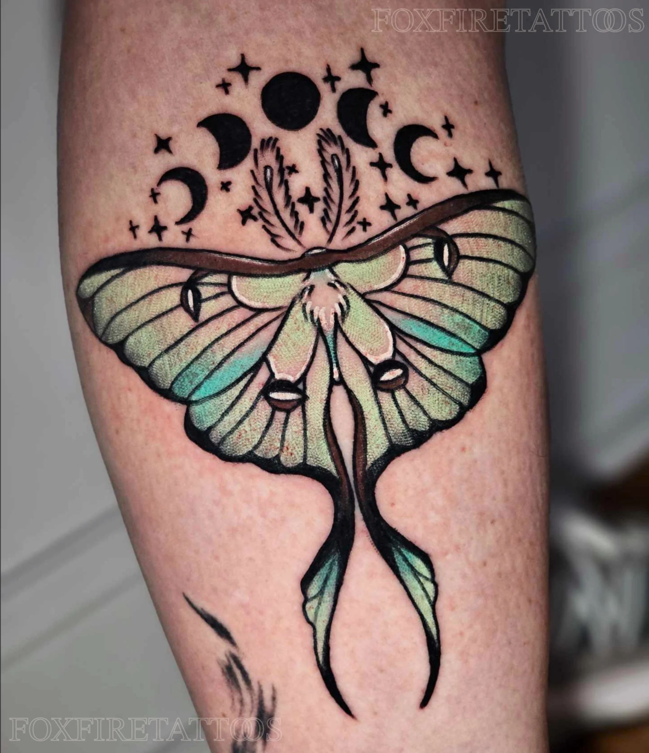 Tattoo of a moth with colorful, detailed wings under a series of moon phases and stars.