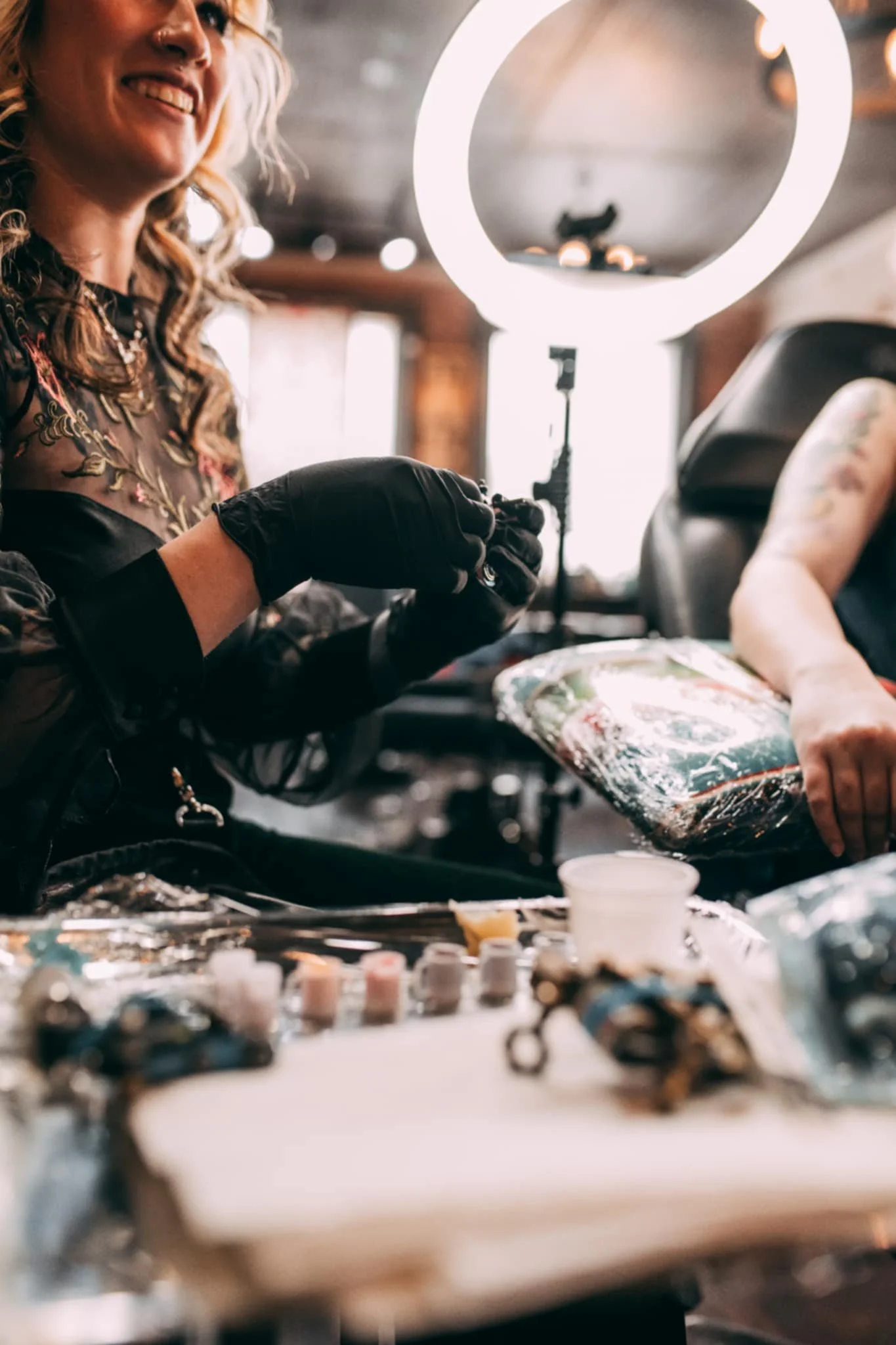 Tattoo artist working at a tattoo station with tattoo supplies on the table, standing in a room with warm lighting and a ring light in the background.