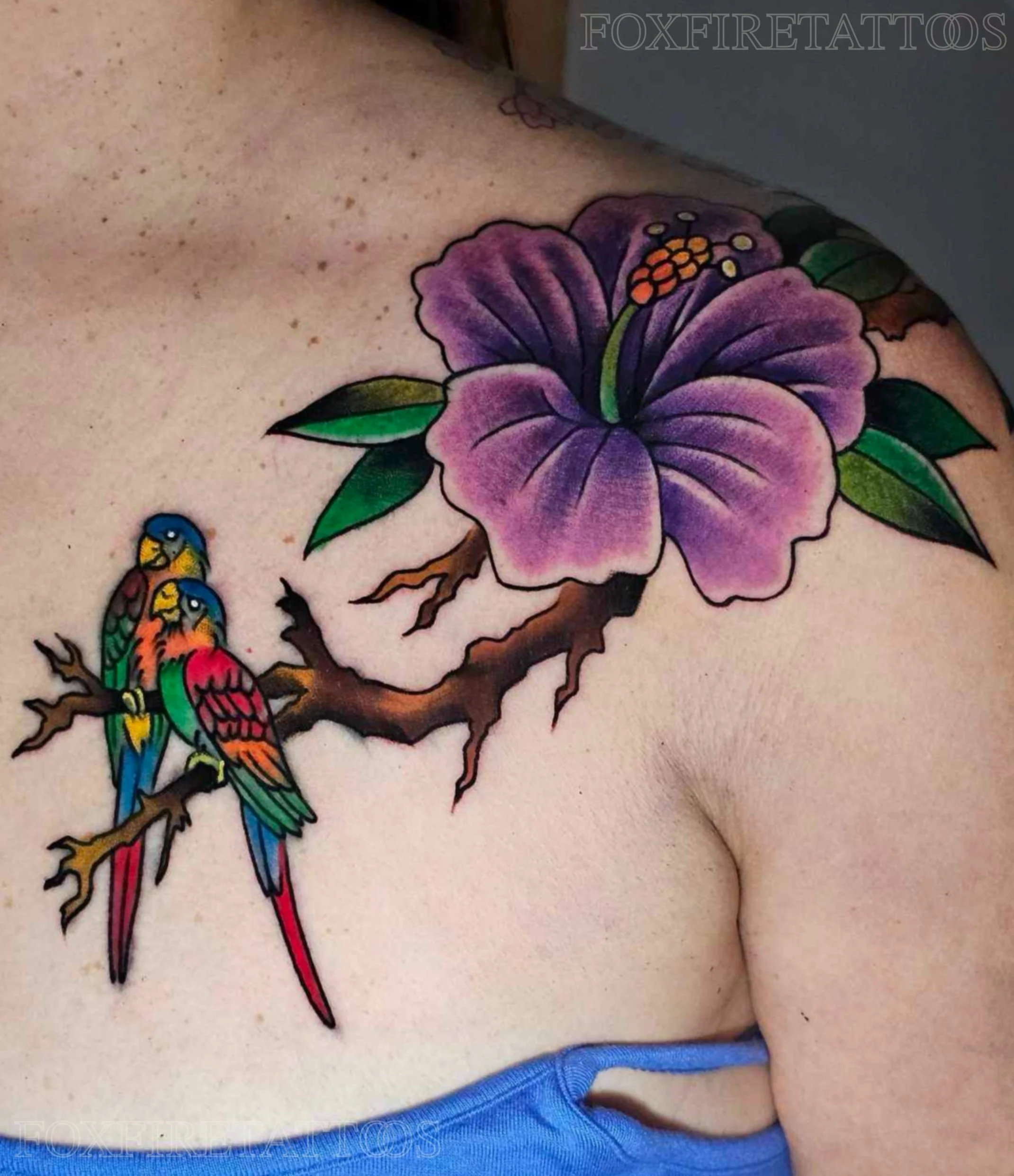 Colorful tattoo of a purple hibiscus flower with green leaves and a brown branch on a person's shoulder, featuring three vibrant parakeets perched on the branch.