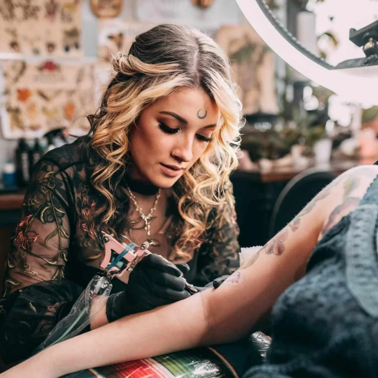 A woman with blonde, curly hair getting a tattoo on her arm in a tattoo studio.