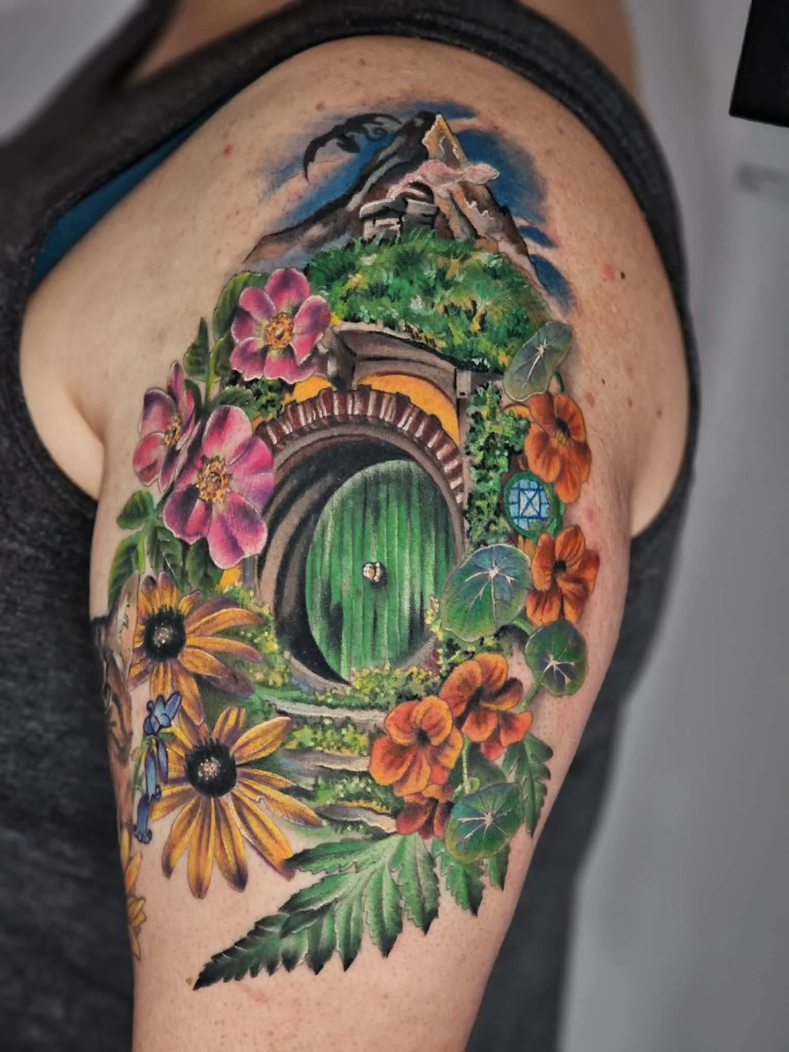 Colorful tattoo on arm featuring a mountain, flowers, a green door, and lush greenery.