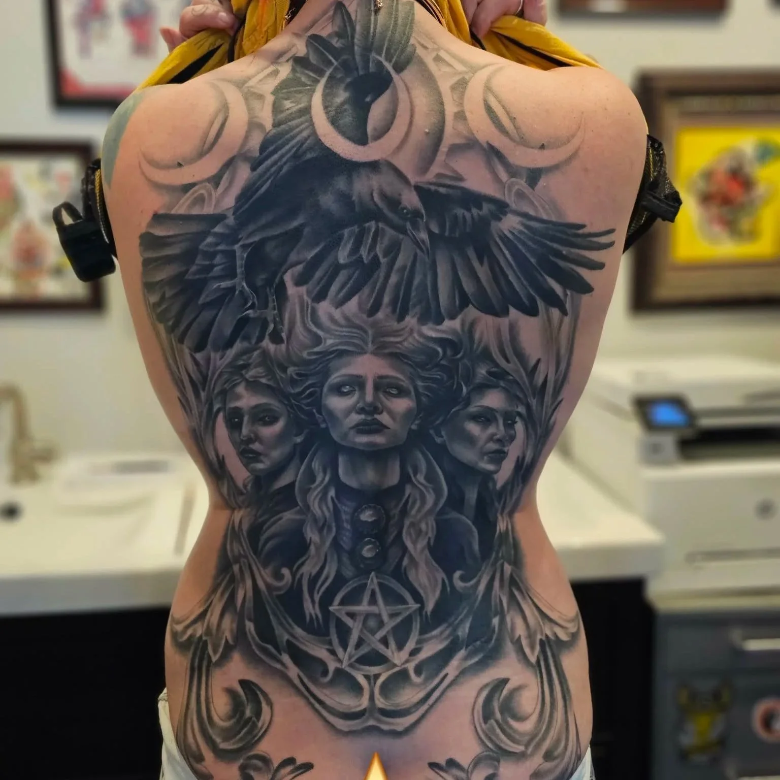 Black and gray tattoo covering woman's back, featuring three women with serious expressions, a large bird with wings spread, and a pentagram symbol.