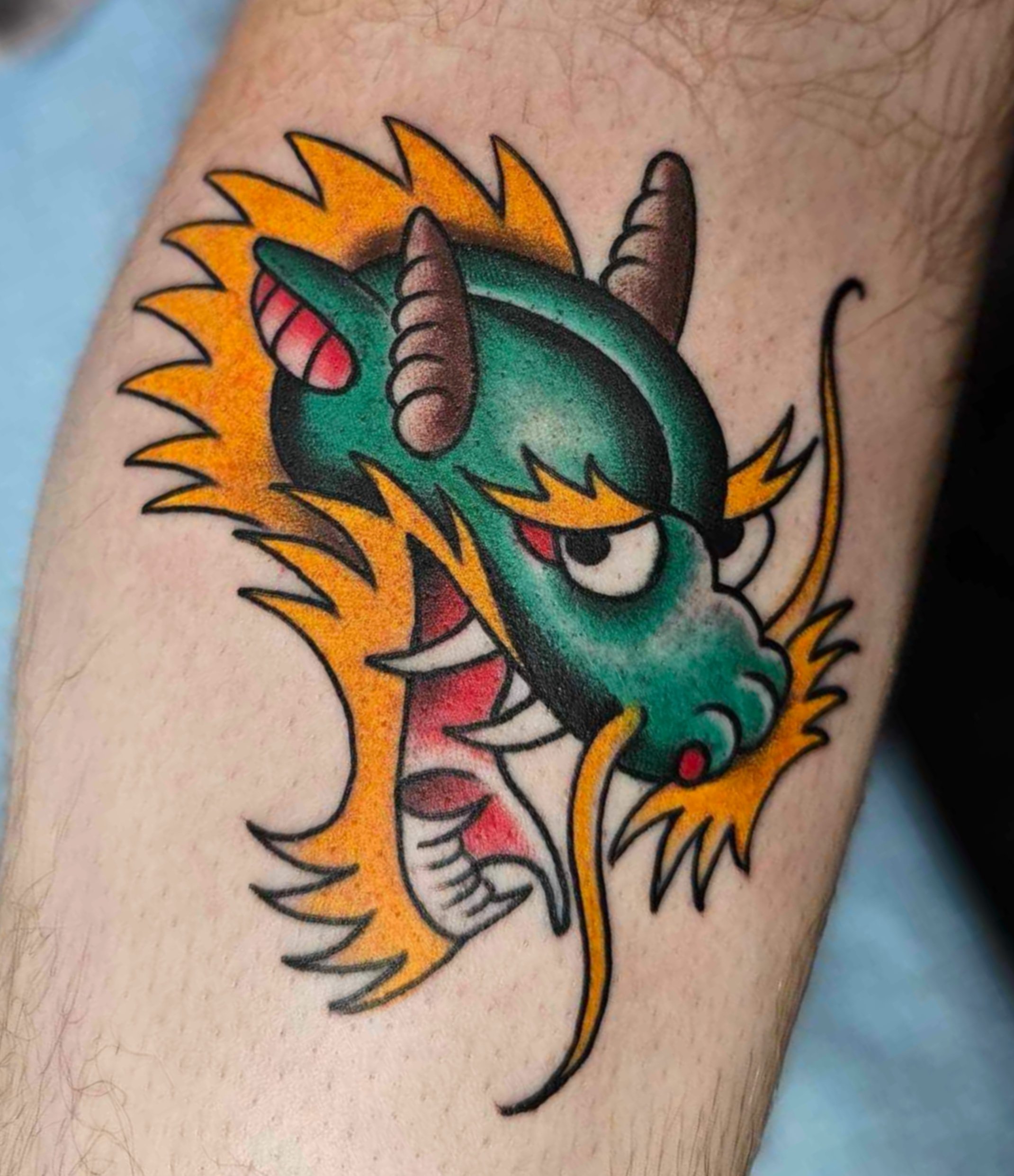 Colorful tattoo of a cartoonish dragon with a green body, orange mane, and horns, featuring a mischievous expression with half-closed eyes.