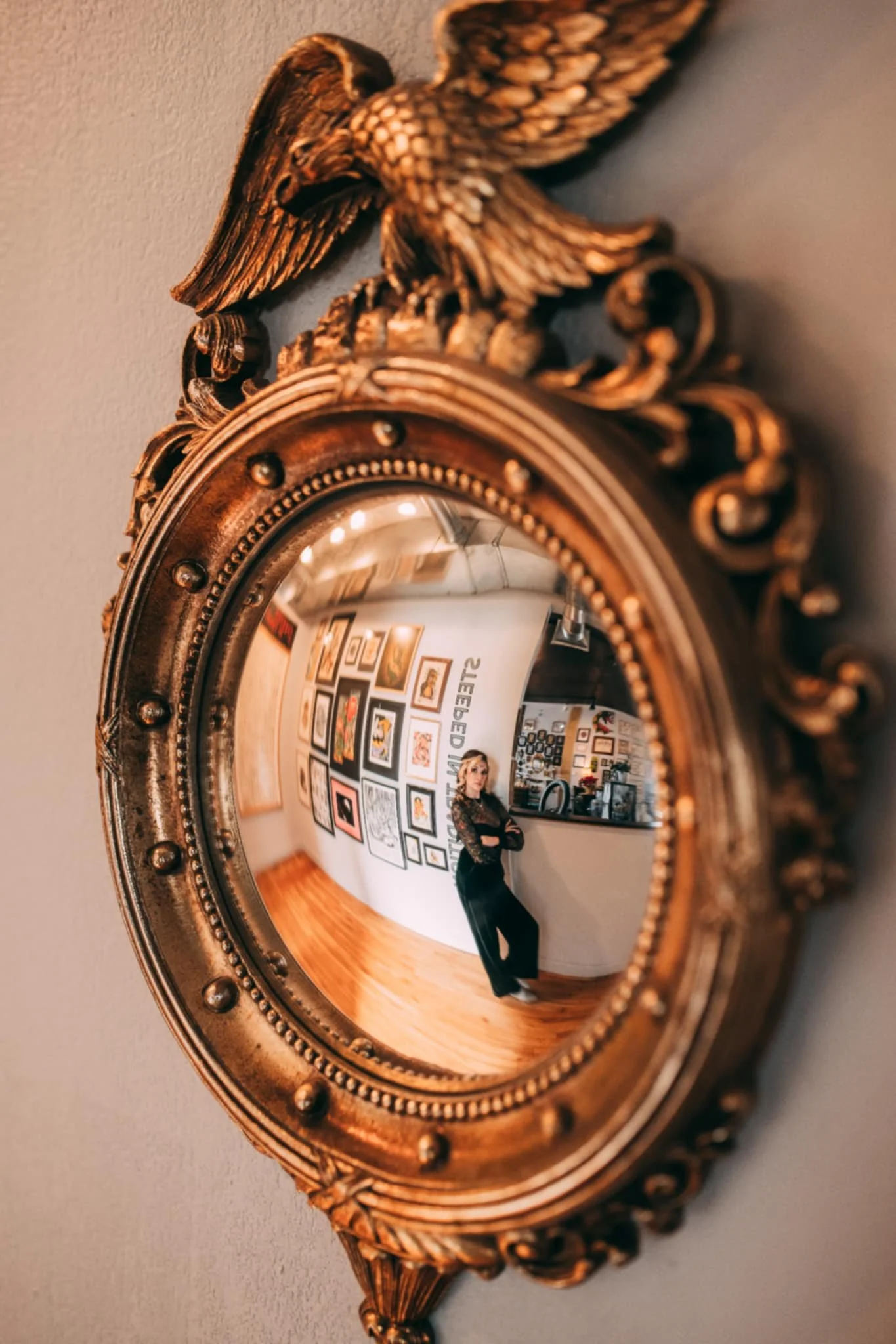 Reflection of woman in a circular, ornate gold mirror hanging on a wall in an art gallery.