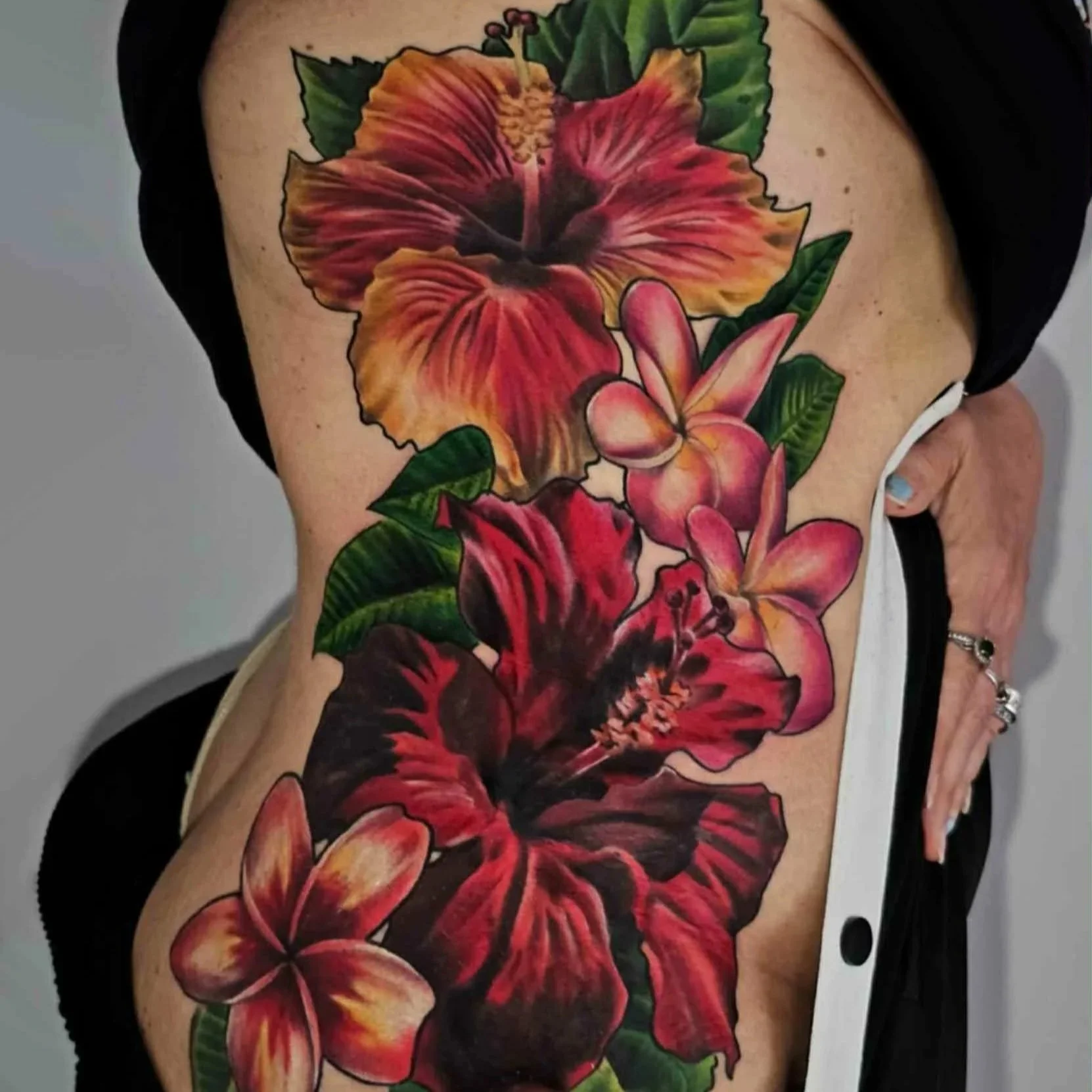 Colorful floral tattoo featuring large hibiscus and plumeria flowers on a person's side torso.