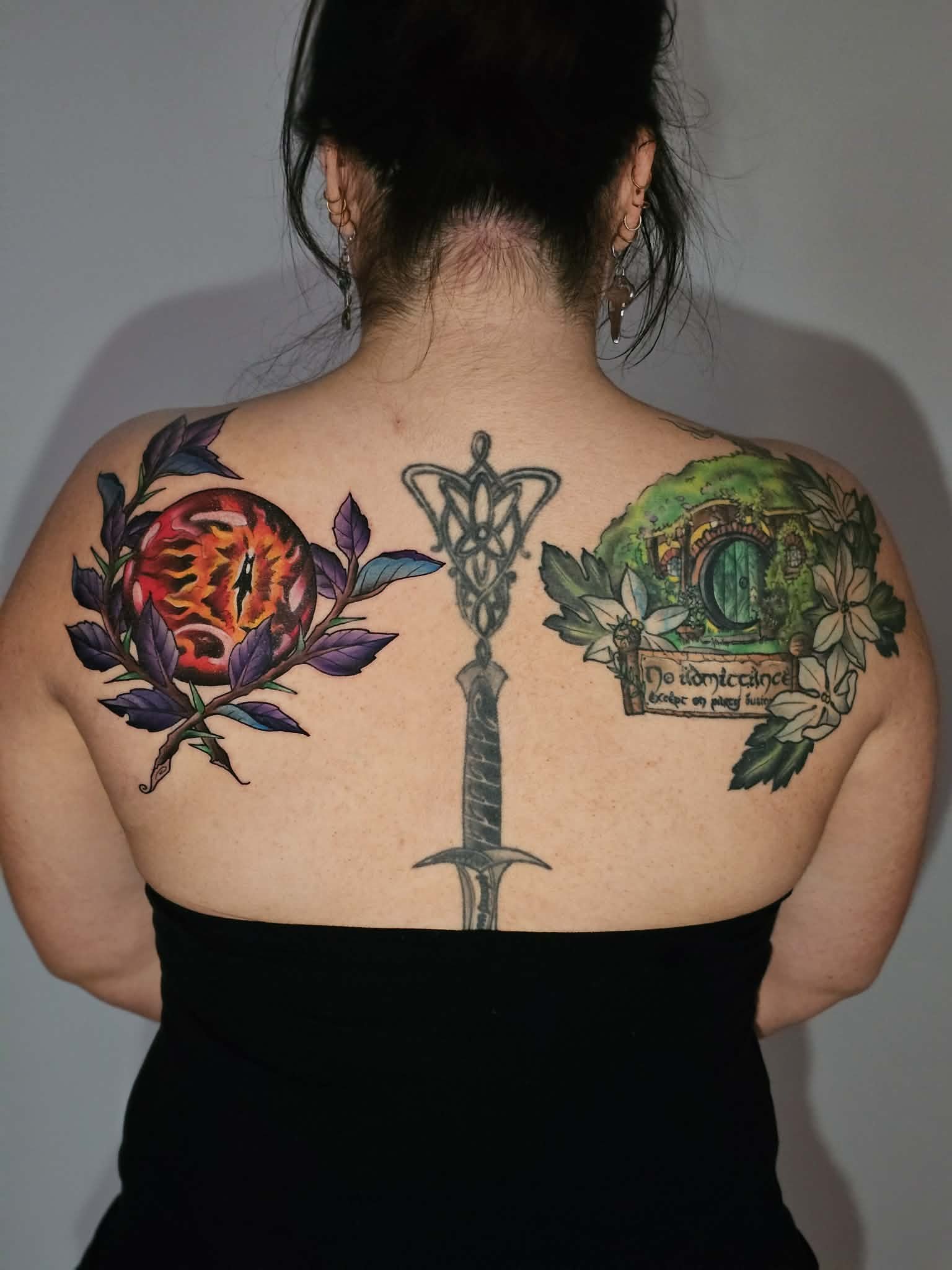 Back of a woman with dark hair tied up, showcasing colorful tattoos on her shoulders and upper back. The tattoos include a flaming eye surrounded by purple leaves on the left, a green door within a garden scene with flowers on the right, and a large 