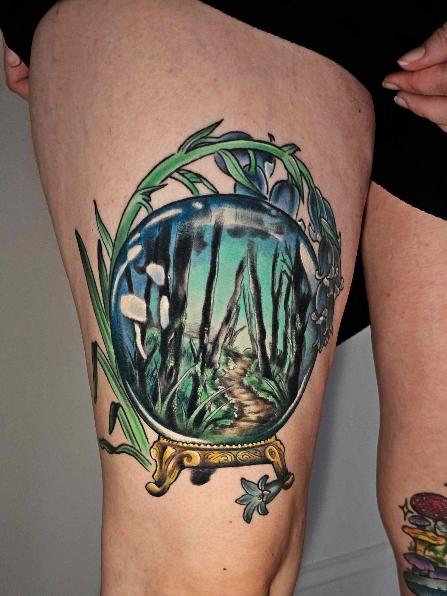 Tattoo of a glass snow globe with a forest scene inside, resting on a decorative gold stand, with green leaves and a small flower around it.