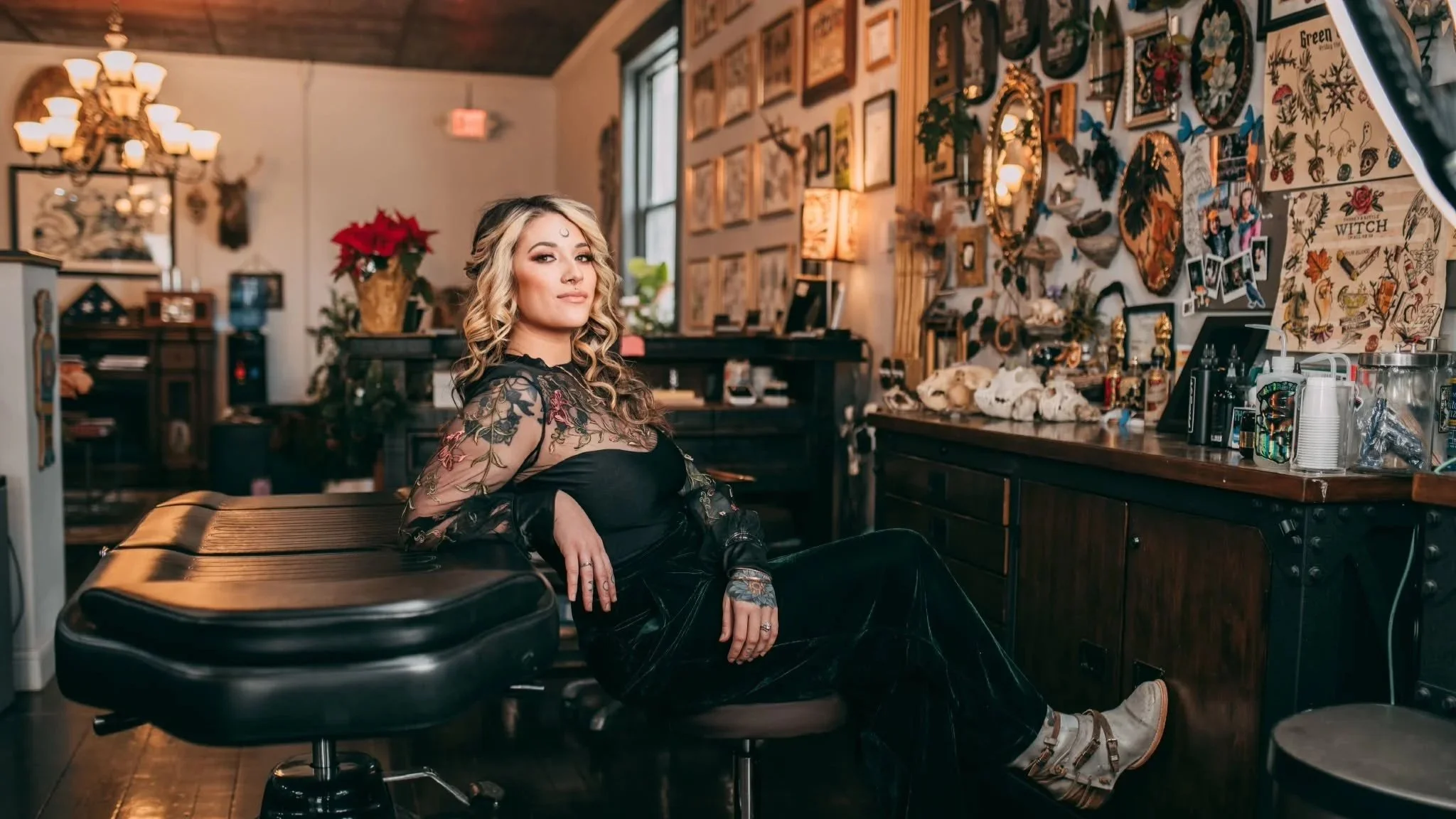 A woman with wavy blonde hair sits in a tattoo studio, surrounded by tattoo designs, skulls, and tattoo equipment on the counter behind her. She is wearing a black lace top with floral embroidery and black pants, looking confidently at the camera.