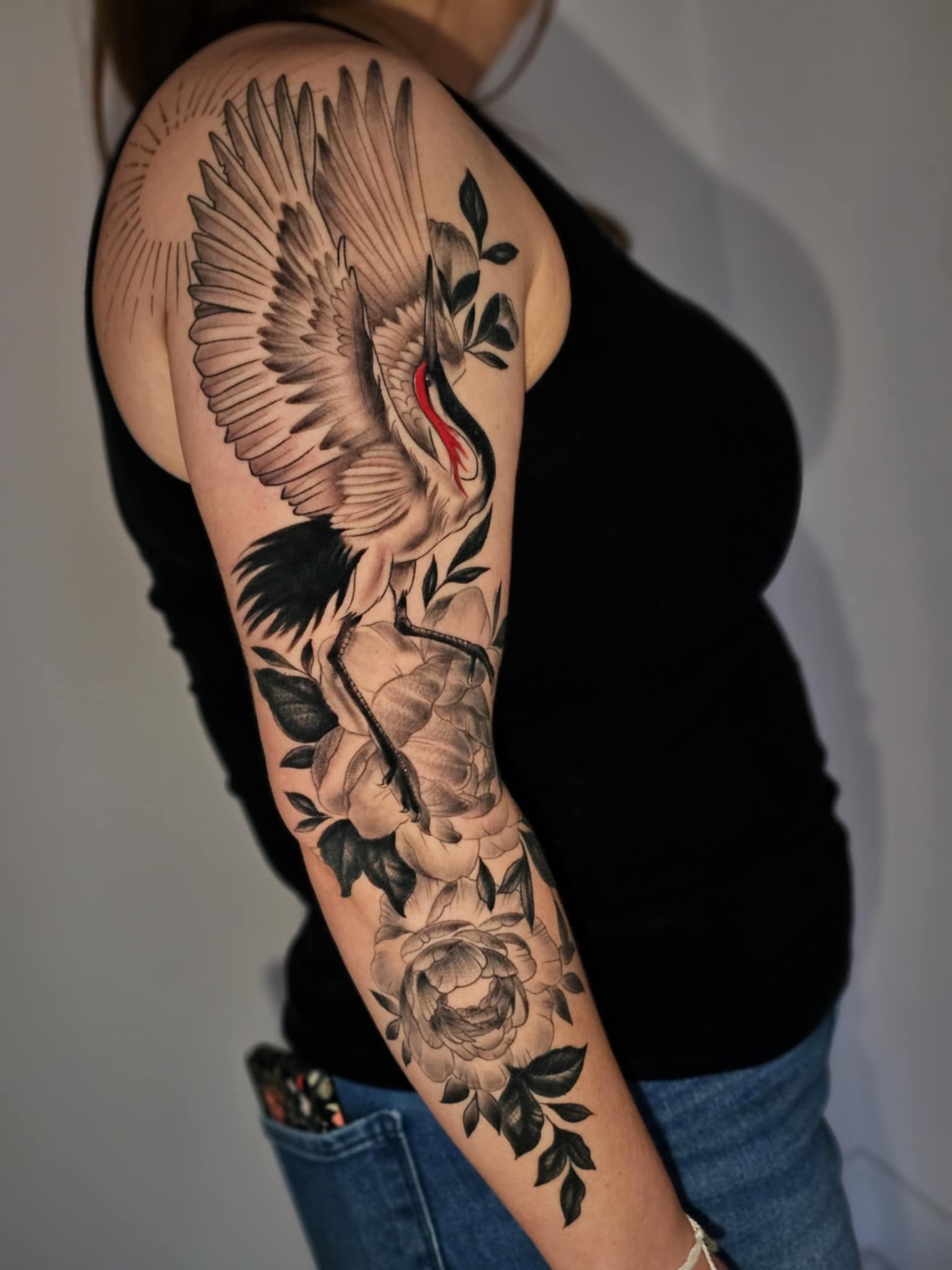 Black and gray tattoo of a crane bird with open wings perched on a branch with flowers and leaves on a woman's arm.