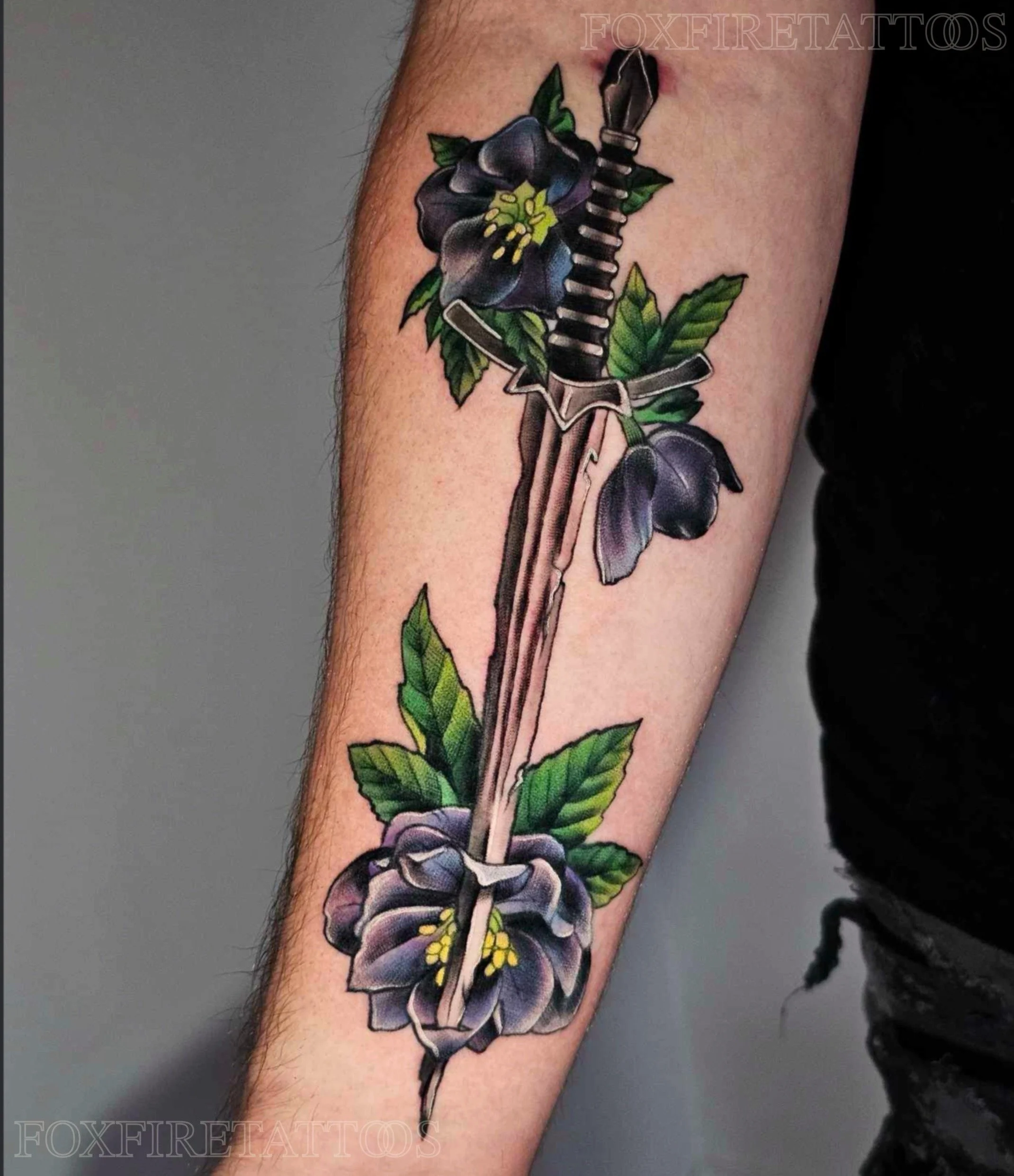 Tattoo of a dagger piercing through purple and black flowers with green leaves on a person's arm.