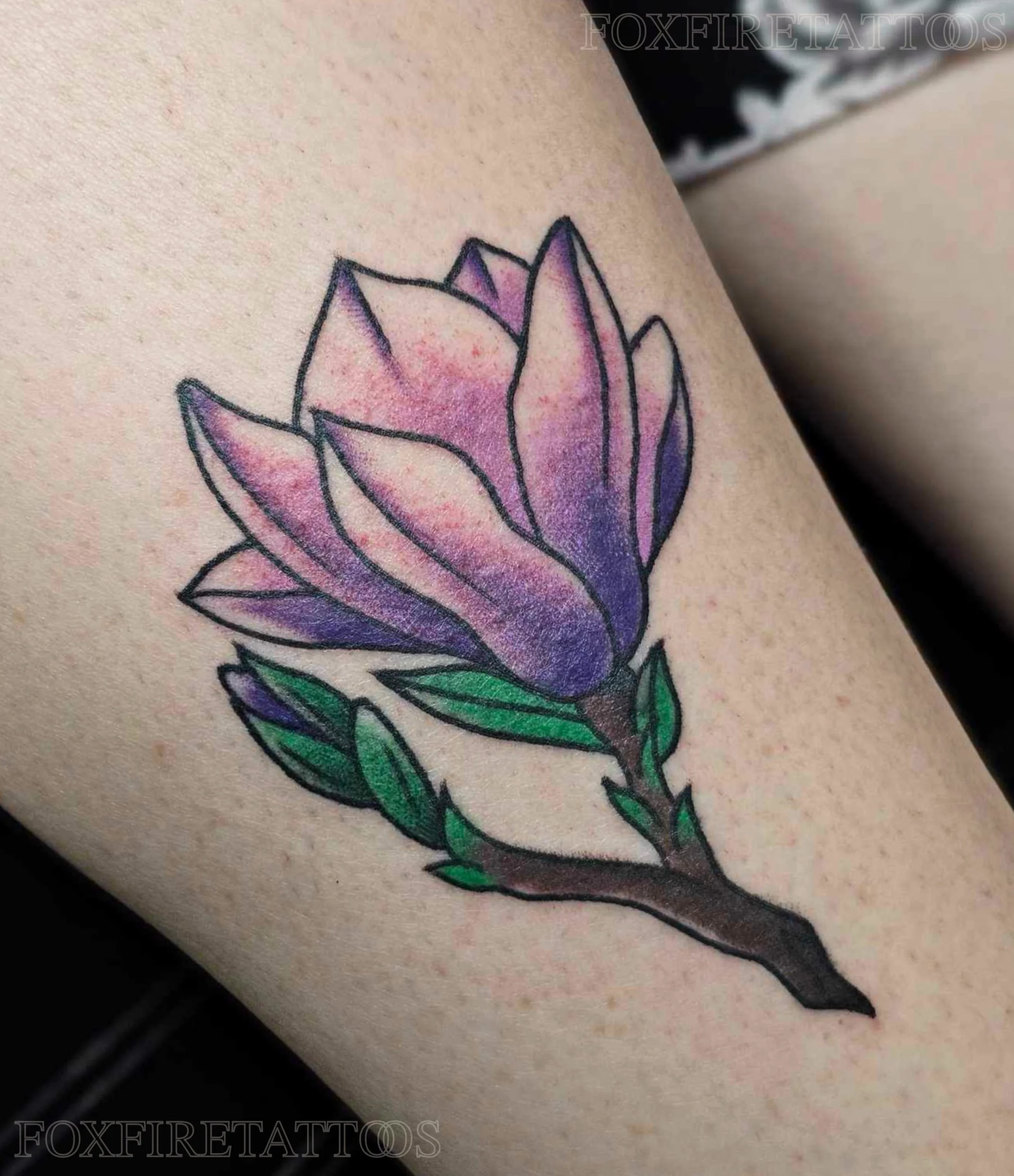 Watercolor style tattoo of a pink and purple magnolia flower with green leaves and dark outline on skin.