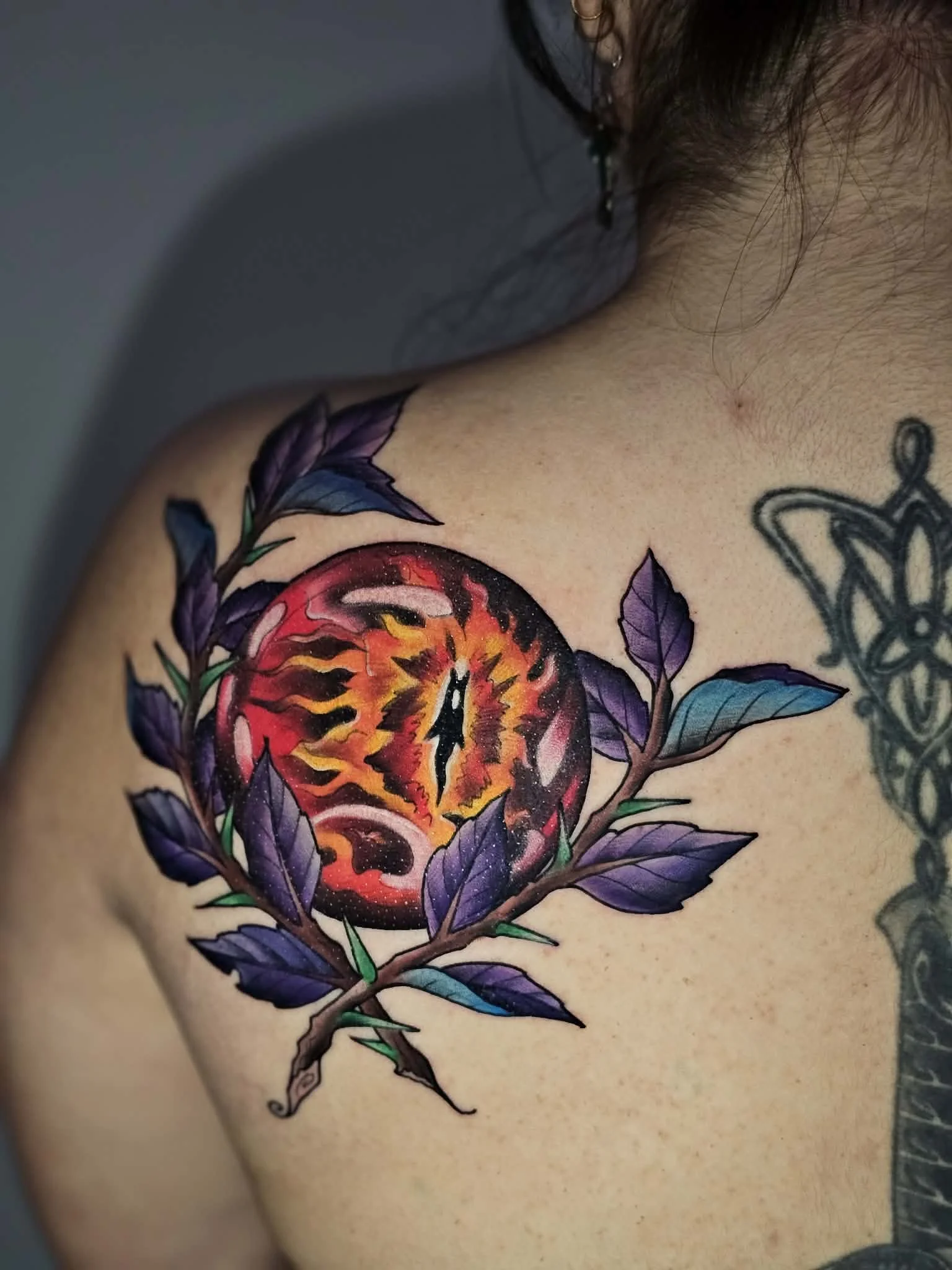 Colorful tattoo of a flaming eye within a black circle, surrounded by purple leaves, on person's shoulder.