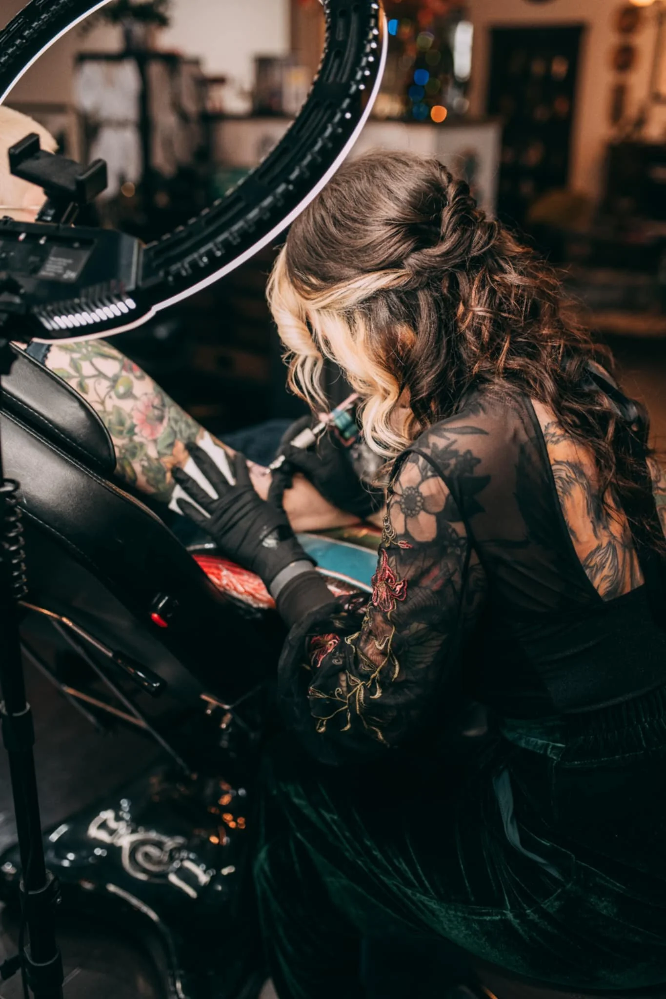 Tattooed woman with styled brown and blonde hair getting a tattoo on her leg in a tattoo studio, with a tattoo machine in hand and tattoo chair visible.