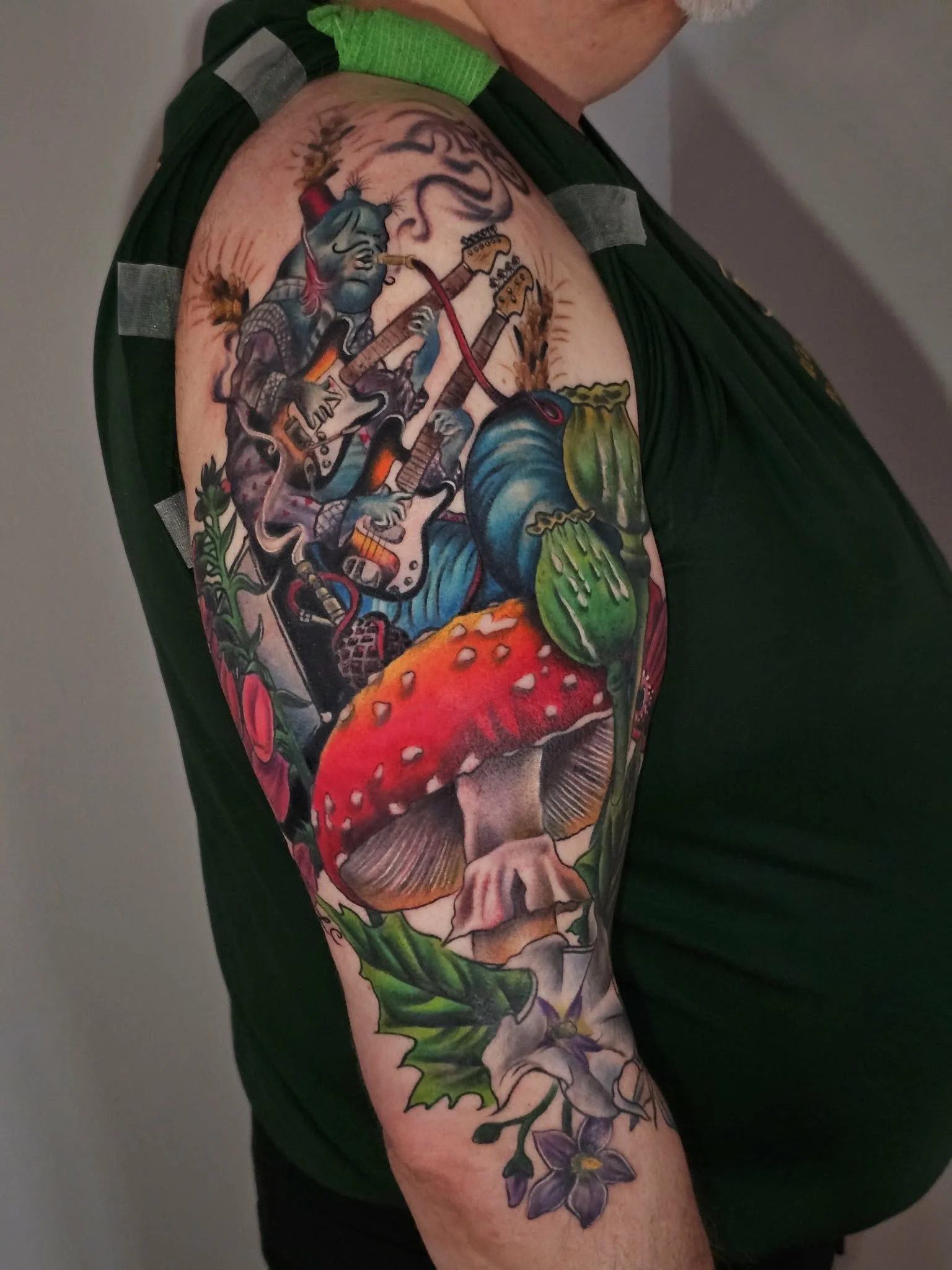 Colorful tattoo sleeve on an upper arm featuring a blue-skinned character with pointed ears playing an electric guitar, surrounded by a mushroom, green leaves, and various decorative elements.