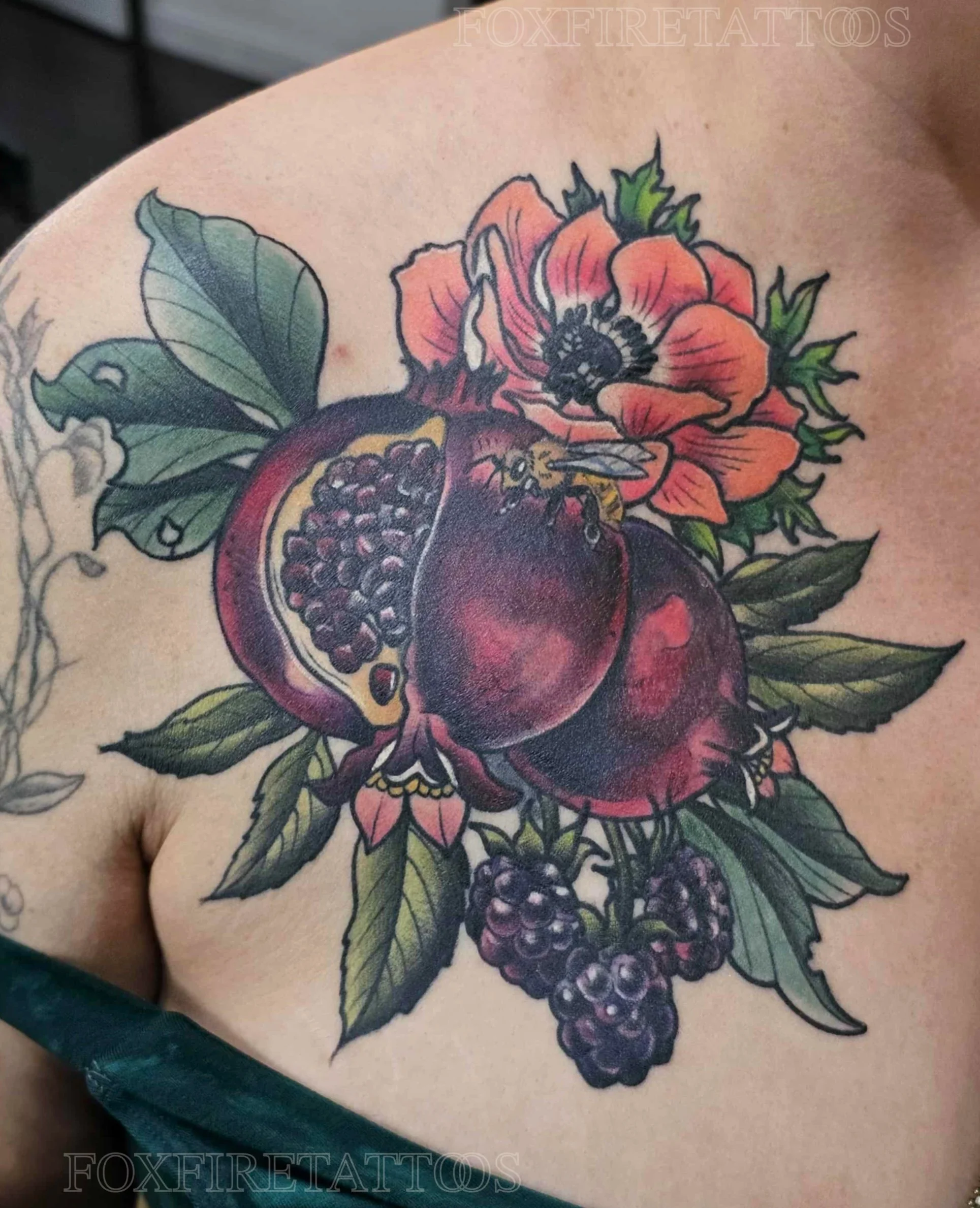 Colorful tattoo of two pomegranates, a bee, a pink flower, and green leaves on a person's shoulder.
