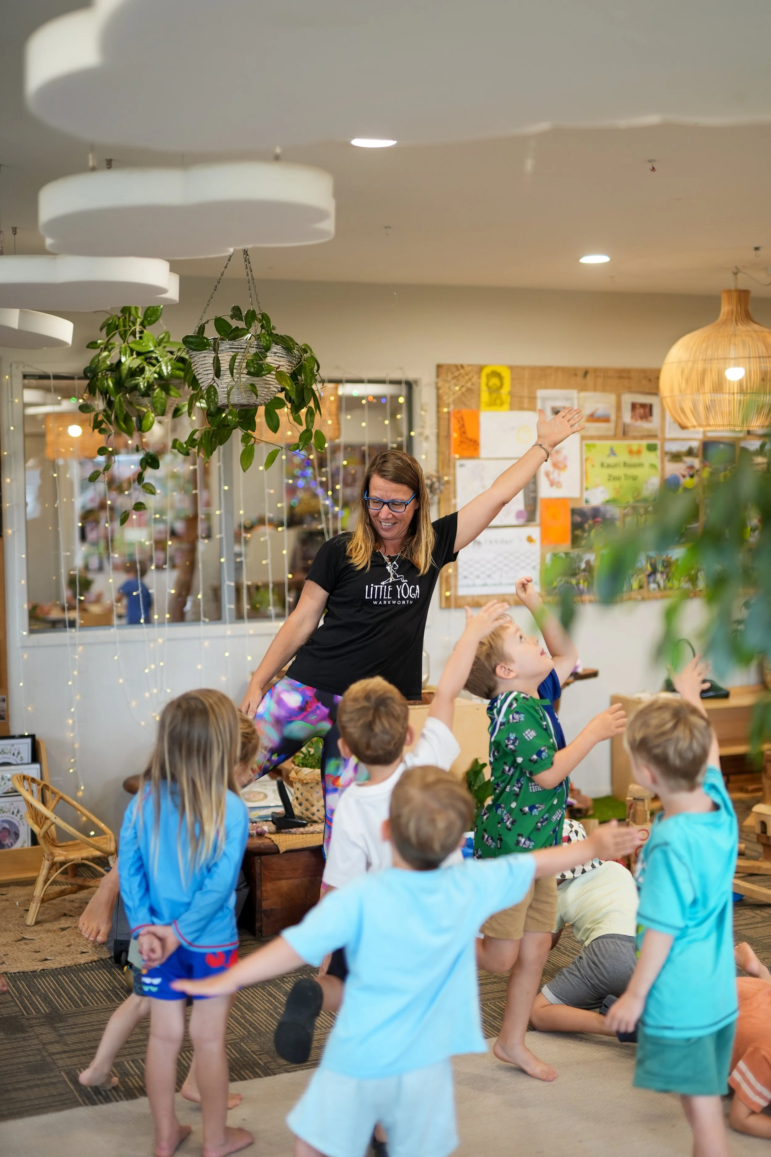 A group of children participating in a yoga class led by an adult instructor in a cozy, decorated room with hanging plants, fairy lights, and bulletin boards.