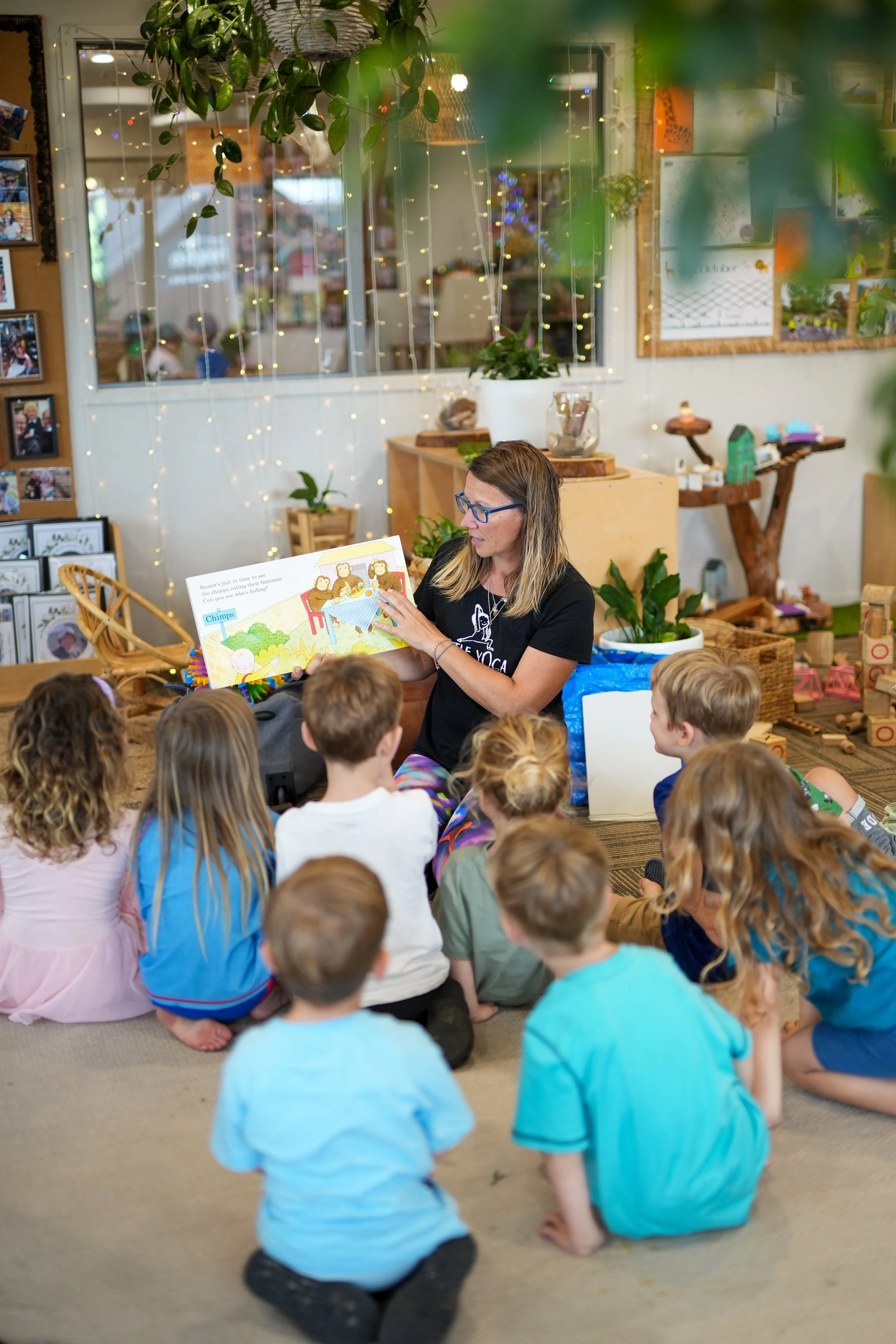 A woman reading a children's book to a group of children sitting on the floor in a cozy indoor space decorated with string lights and plants.