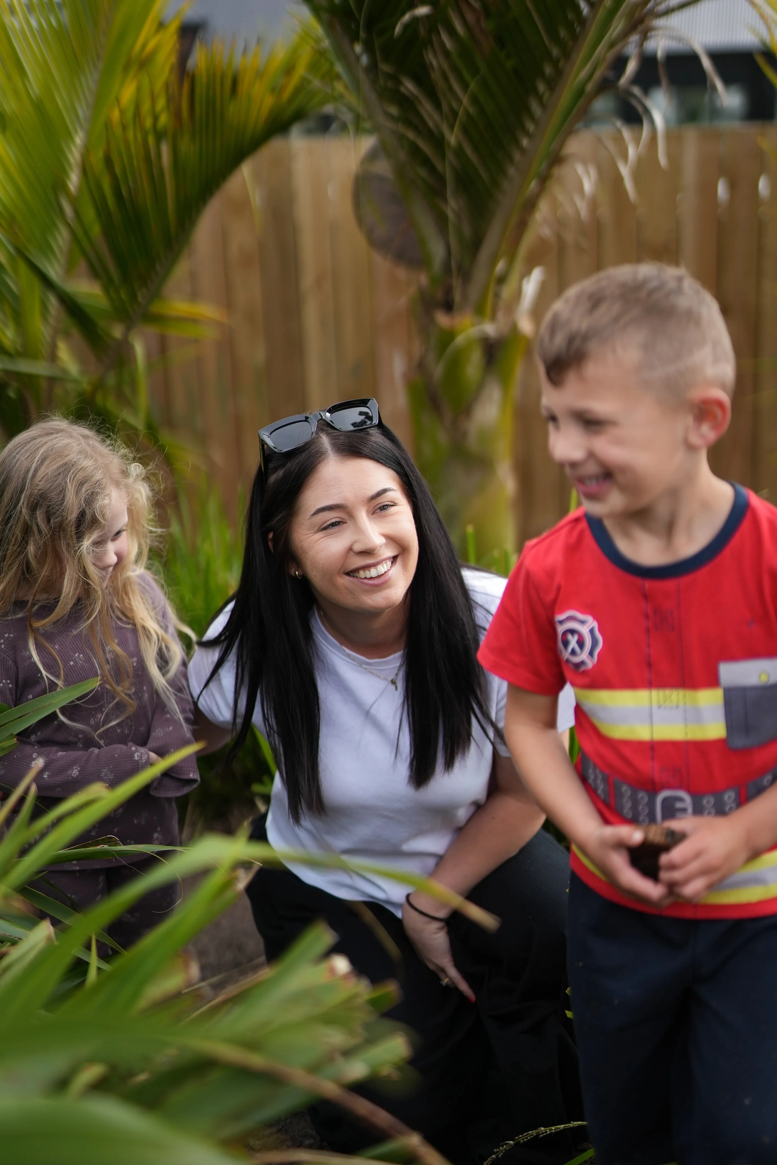 A woman with long dark hair, wearing sunglasses on her head and a white shirt, smiling and talking to two children in a garden with green plants and a wooden fence in the background.