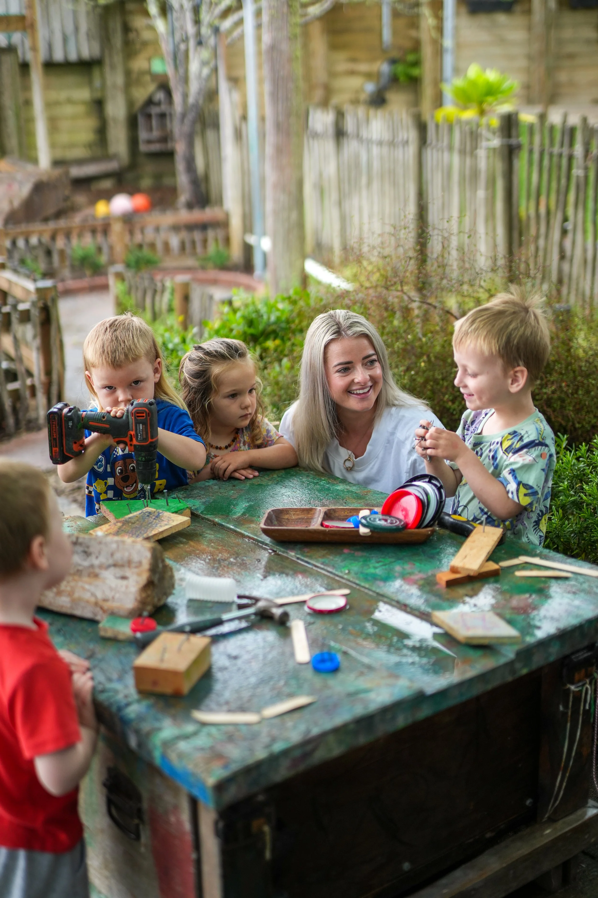 A woman and five children around a weathered green outdoor workbench in a backyard, engaging in a science or crafting activity with various tools and materials on the table.
