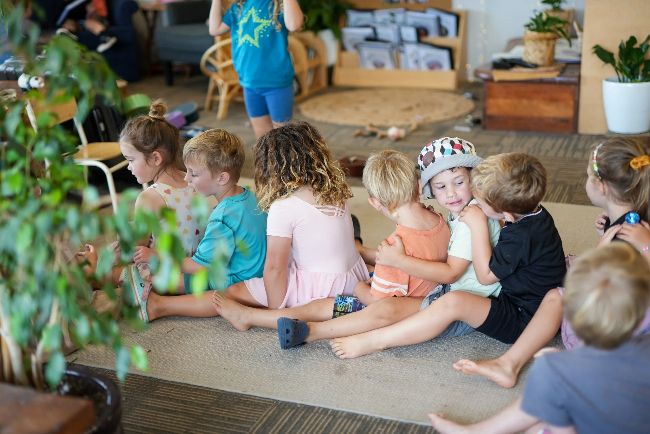 Children sitting on the floor in a line, engaged in a similar activity, in a cozy indoor setting with plants and furniture.