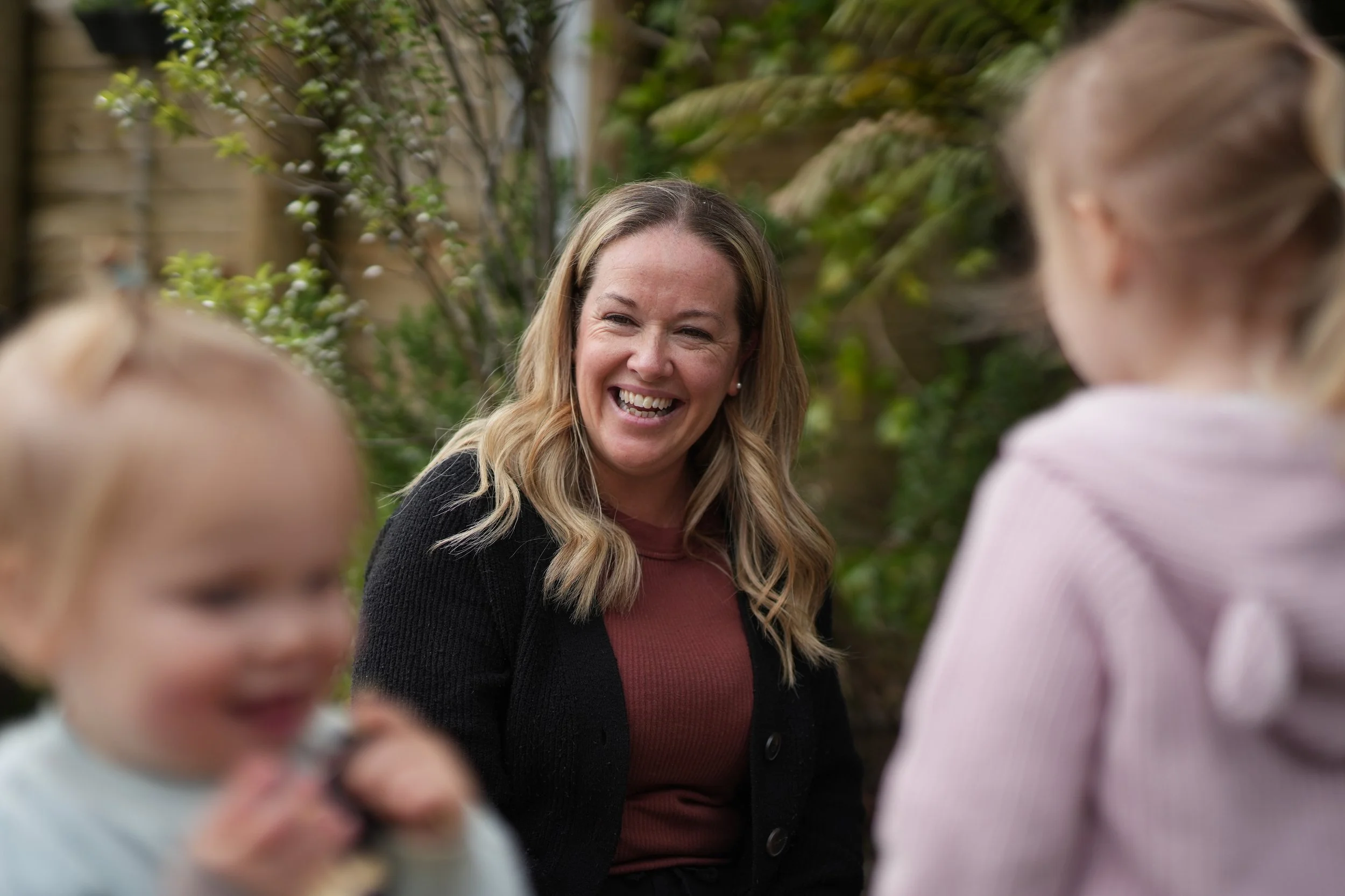A woman with long blonde hair laughing with two young girls outdoors amidst greenery.