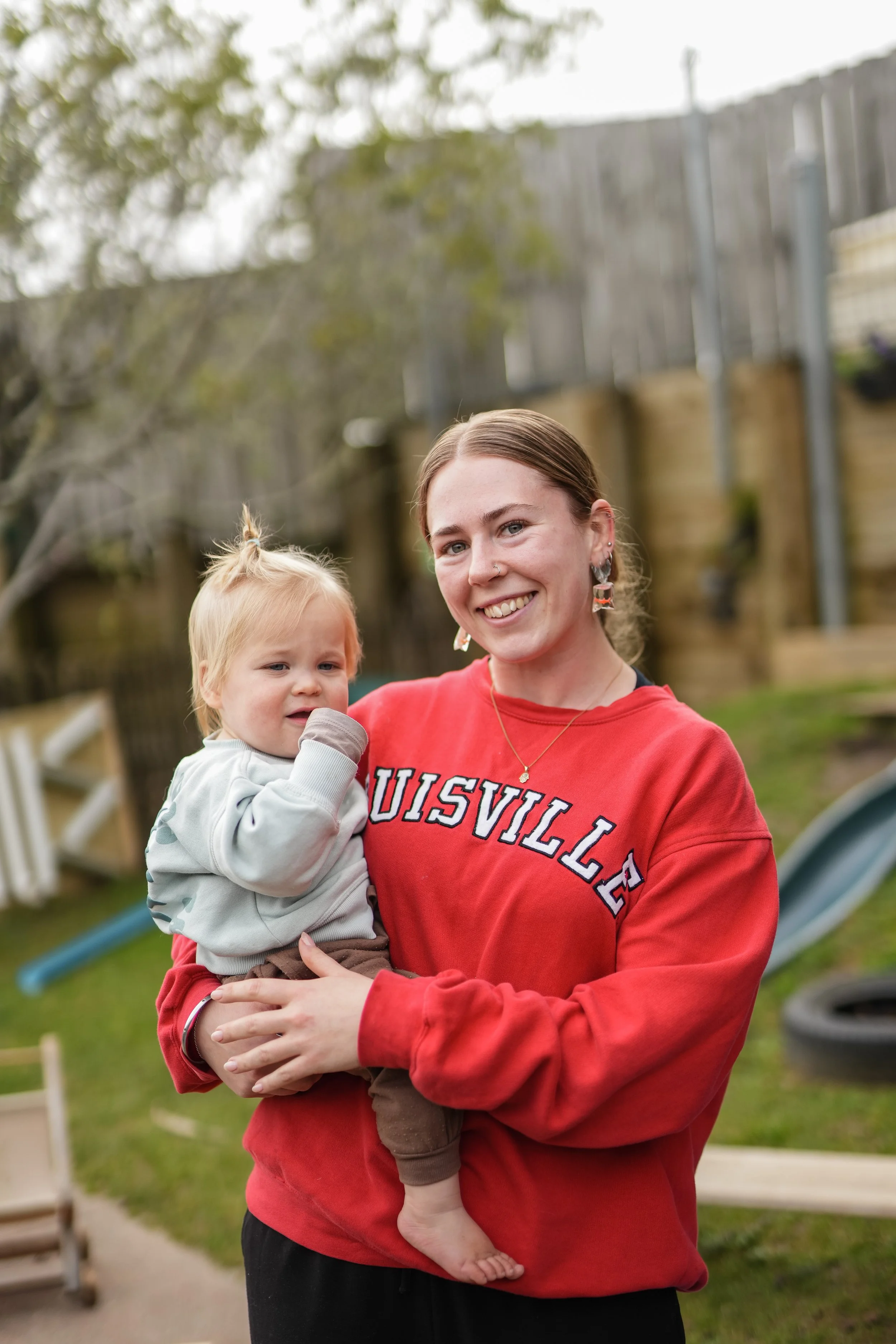 A woman holding a young child in a backyard with green grass, a slide, tires, and wooden fencing, smiling at the camera.