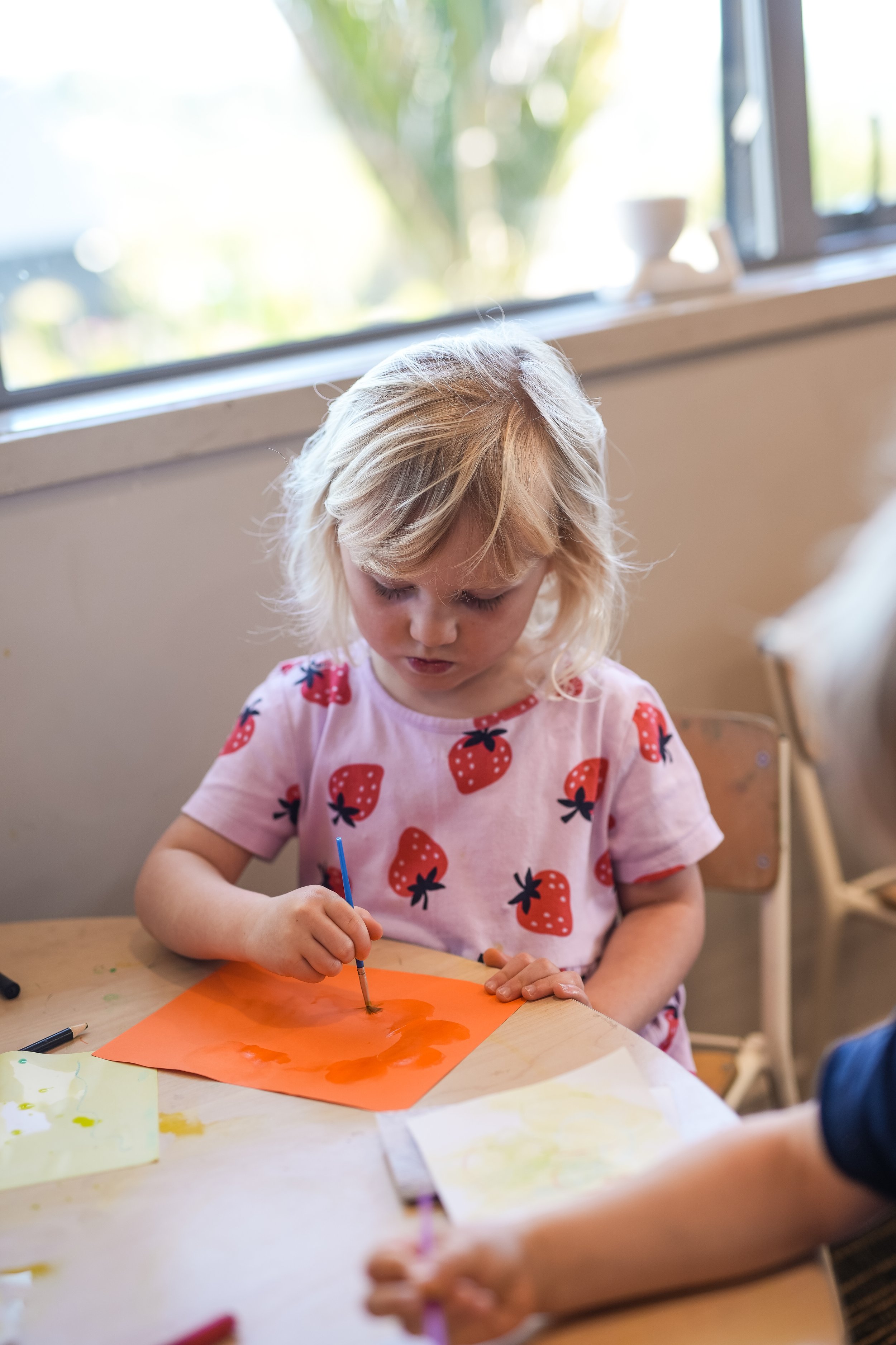 A young girl with blonde hair painting on an orange sheet of paper with a small paintbrush, sitting at a table.