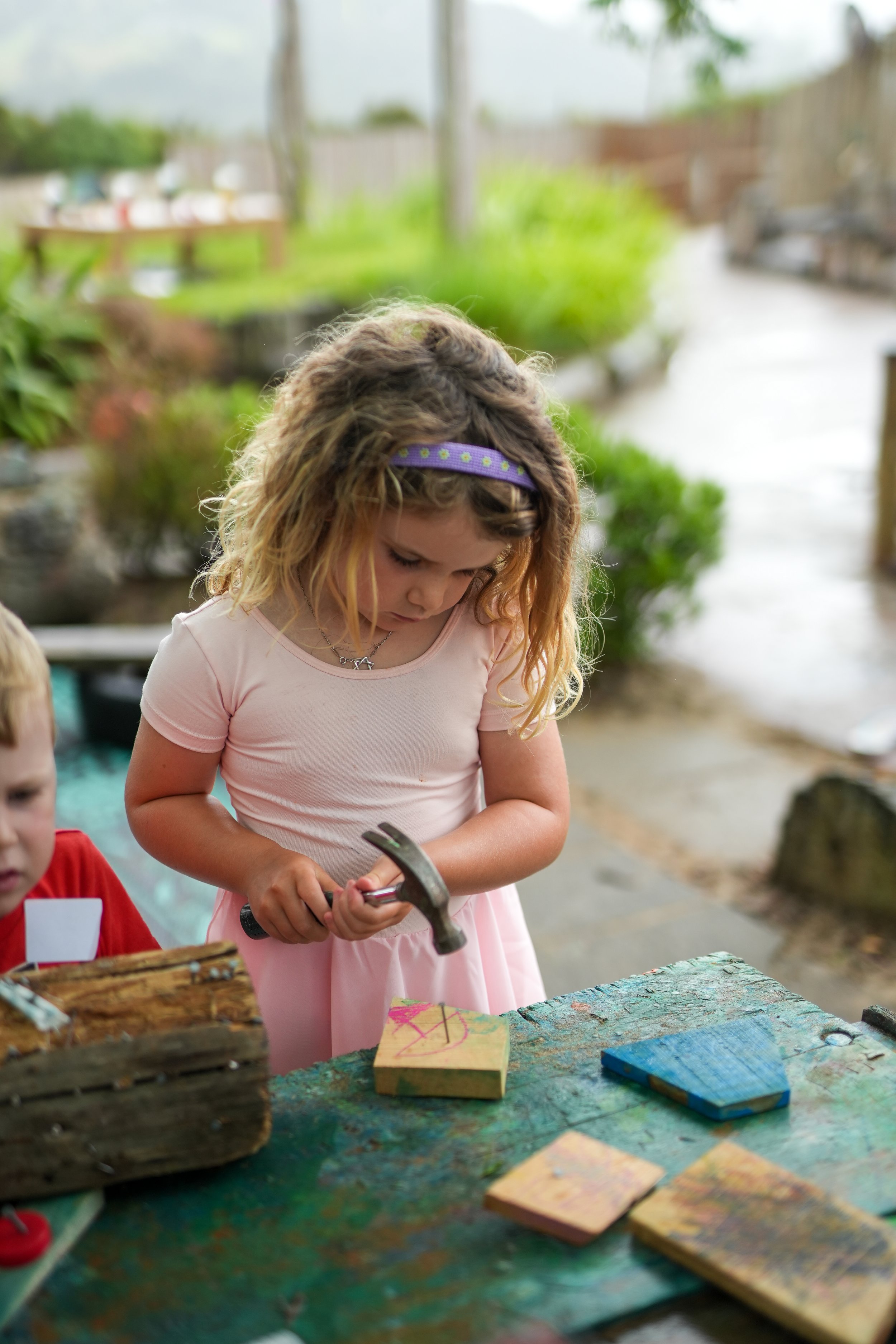 Young girl with curly hair wearing a pink shirt and purple headband hammering a wooden block on a worktable outdoors.