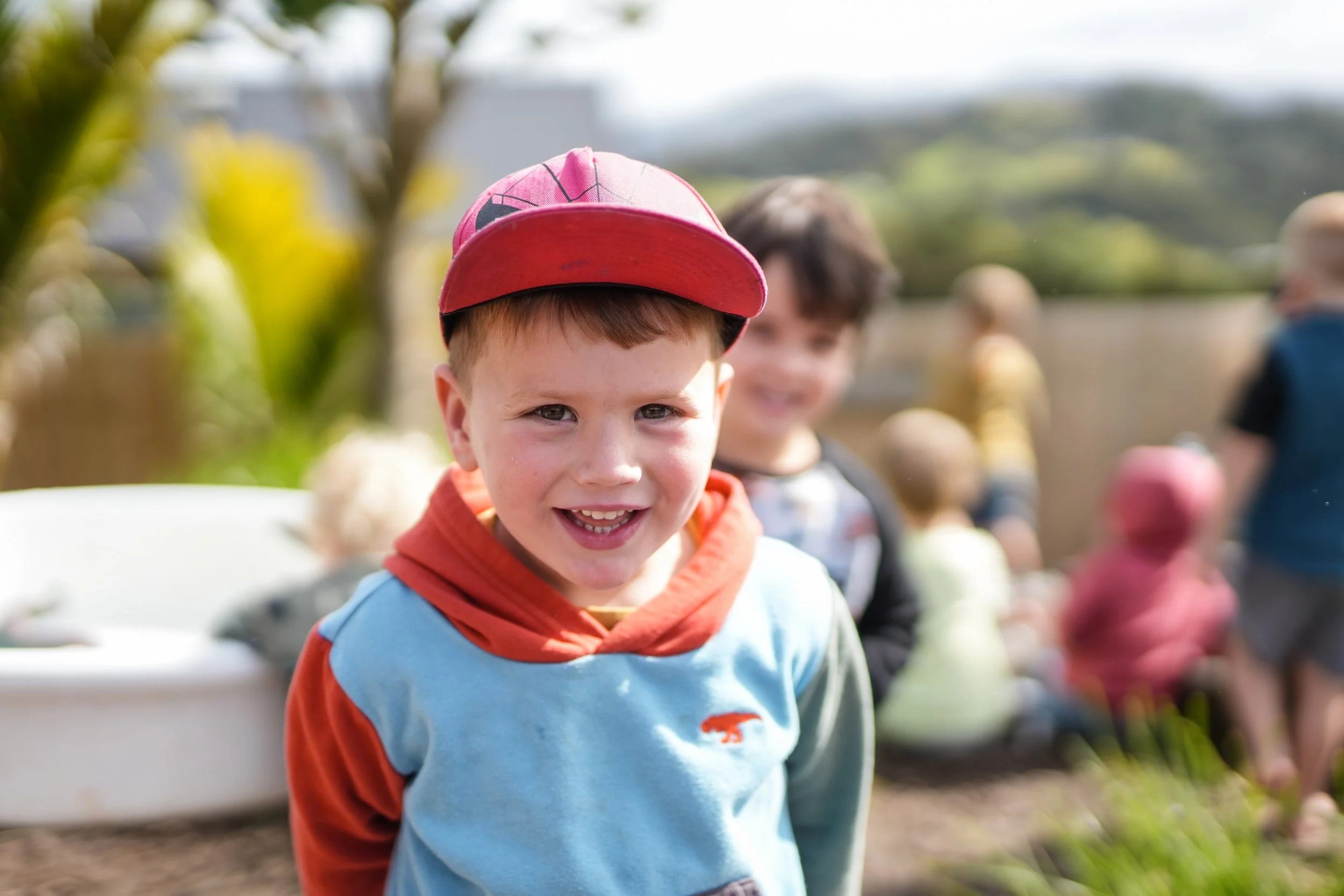 A young boy with a red cap and a blue and orange hoodie smiling at the camera outdoors, with other children in the background.