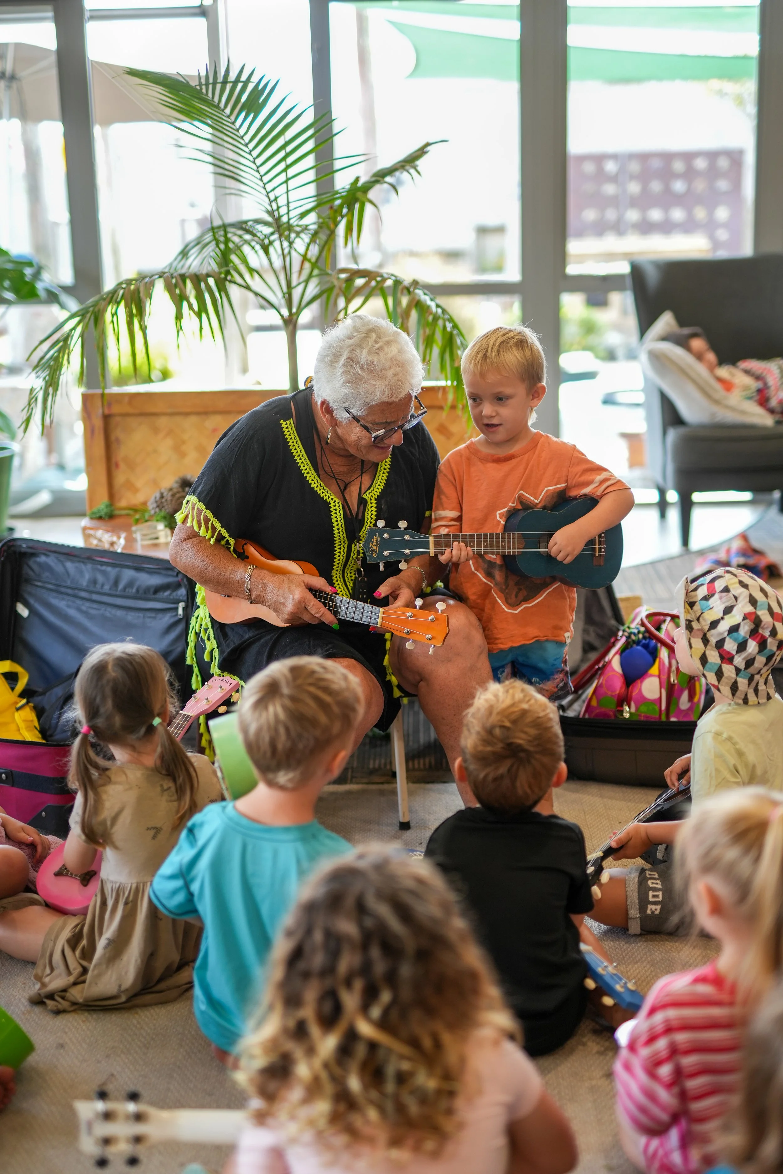 An elderly woman playing a ukulele surrounded by children, some holding small guitars, in a brightly lit indoor setting with large windows and plants.