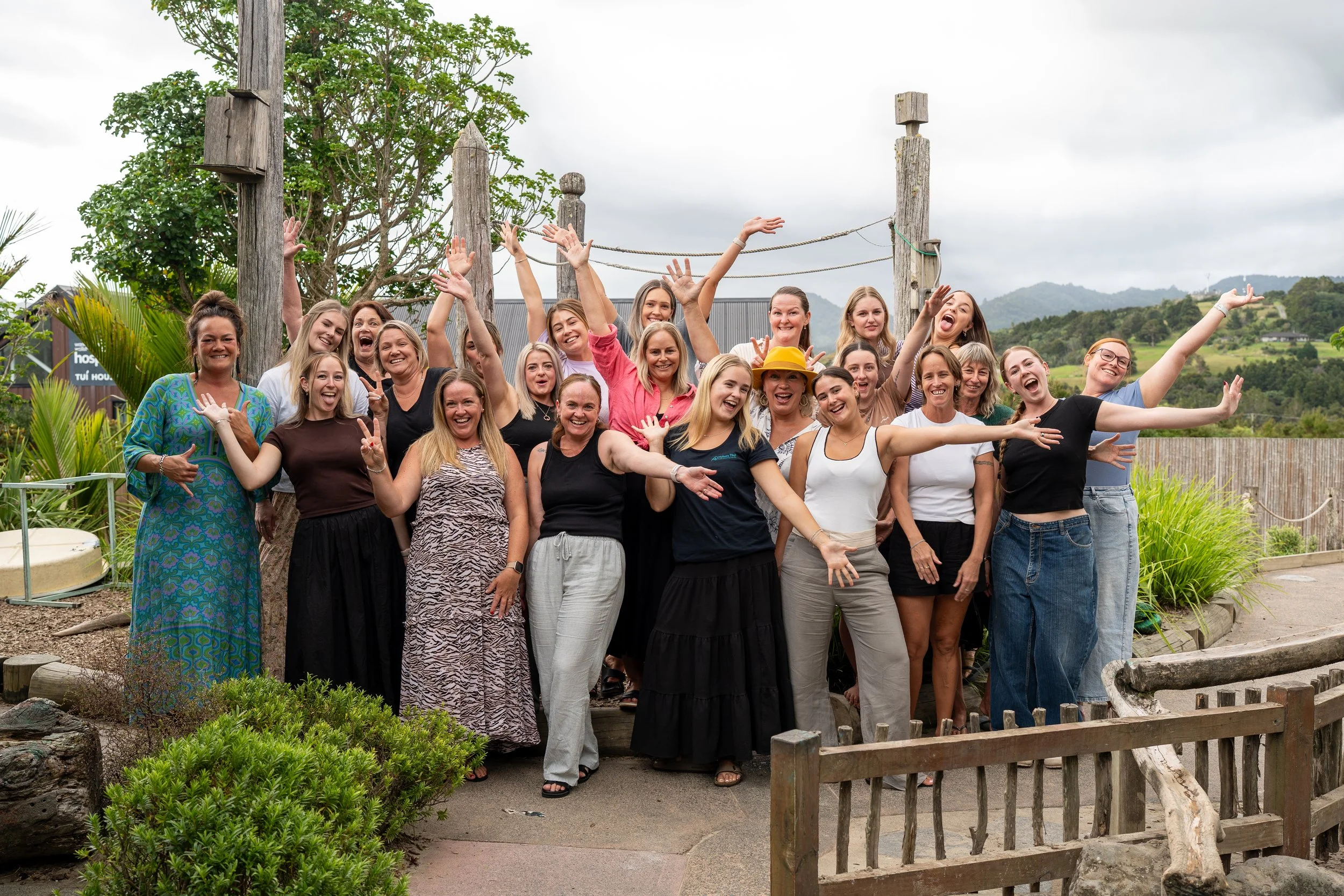 Group of women posing outdoors, some making peace signs, smiling, and gesturing happily, with greenery and hills in the background.