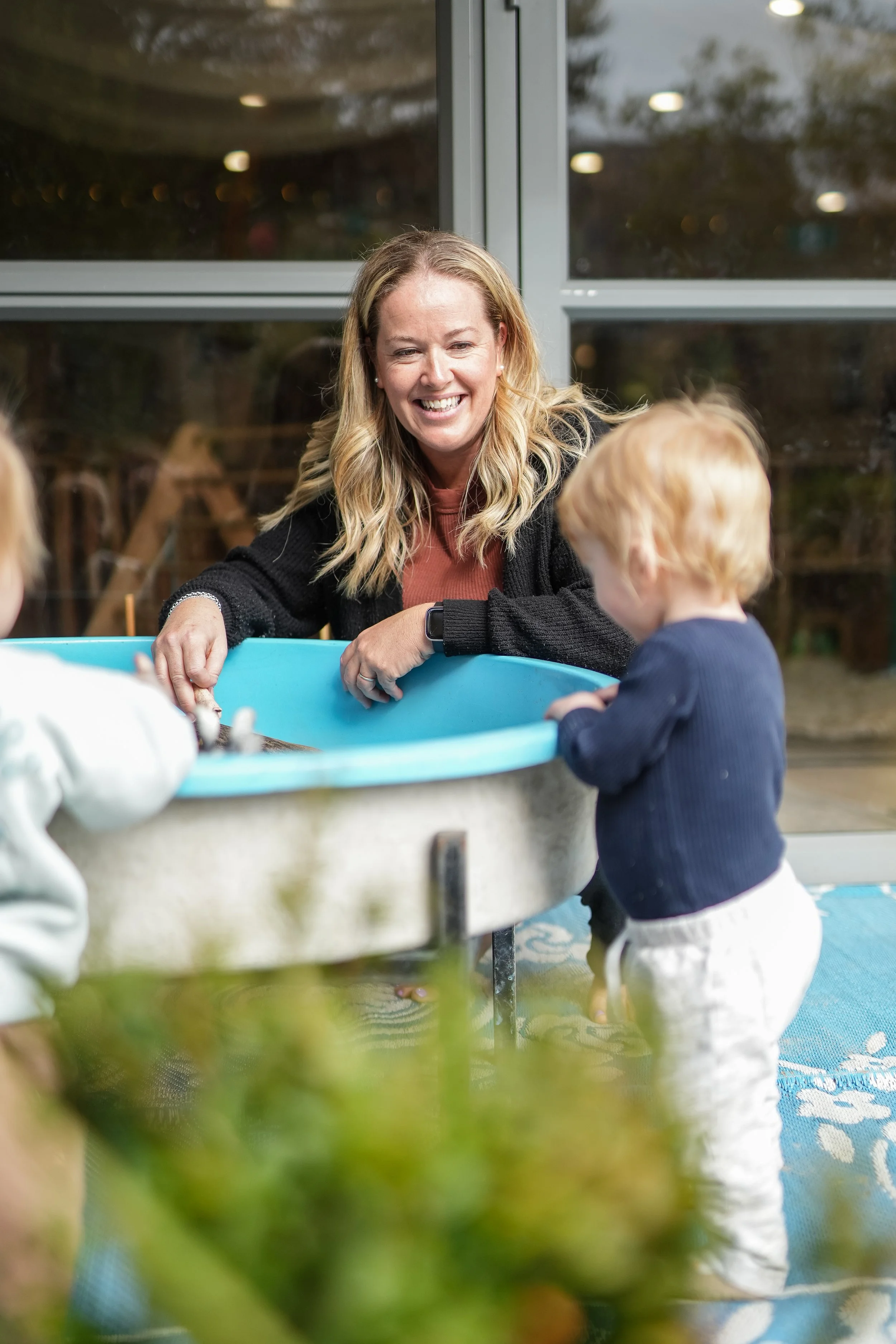 A woman with blonde hair smiling while engaging with young children at an indoor water table.