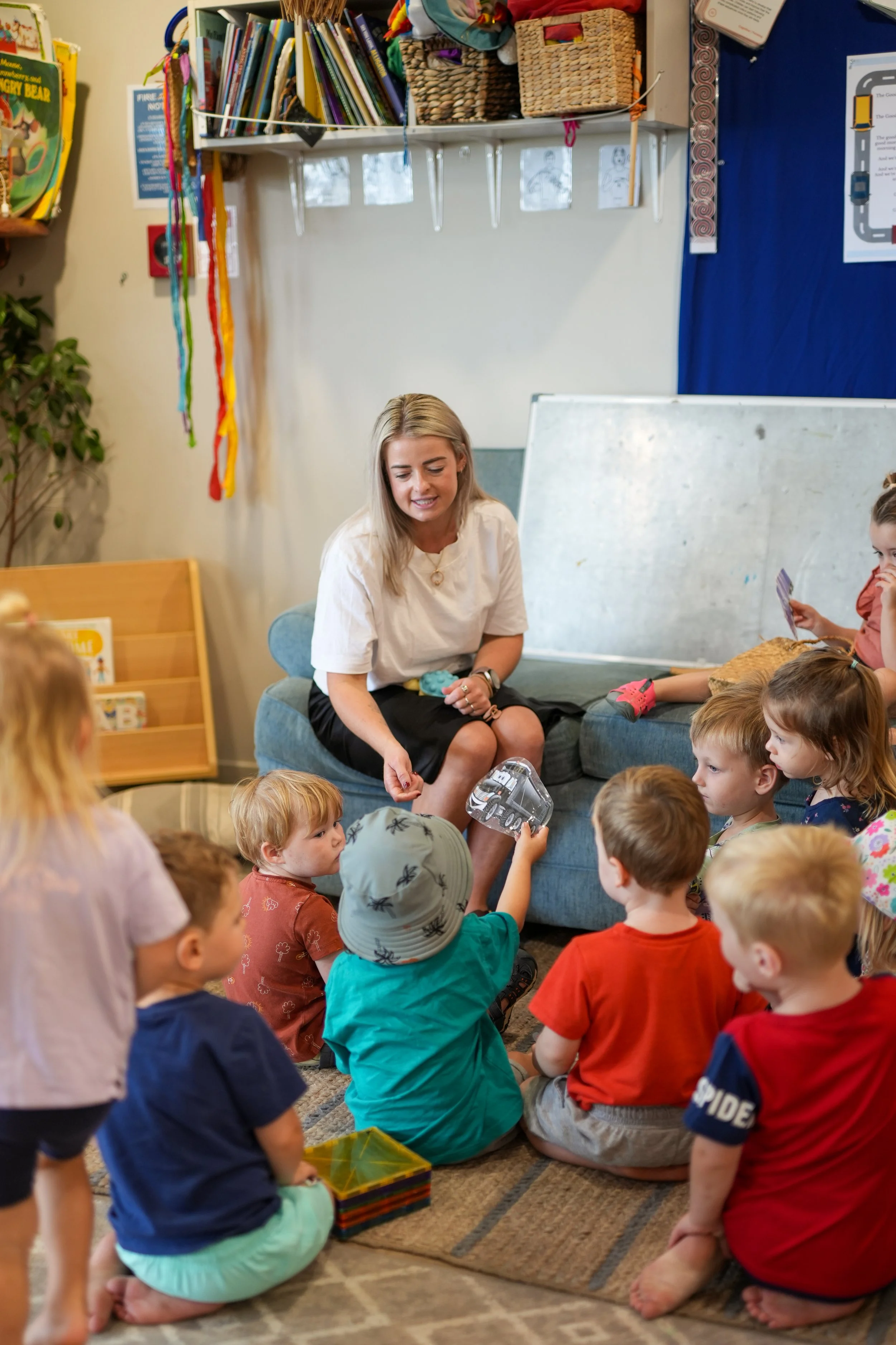 A teacher reading to a group of young children in a classroom, sitting on a rug with books and educational materials around.