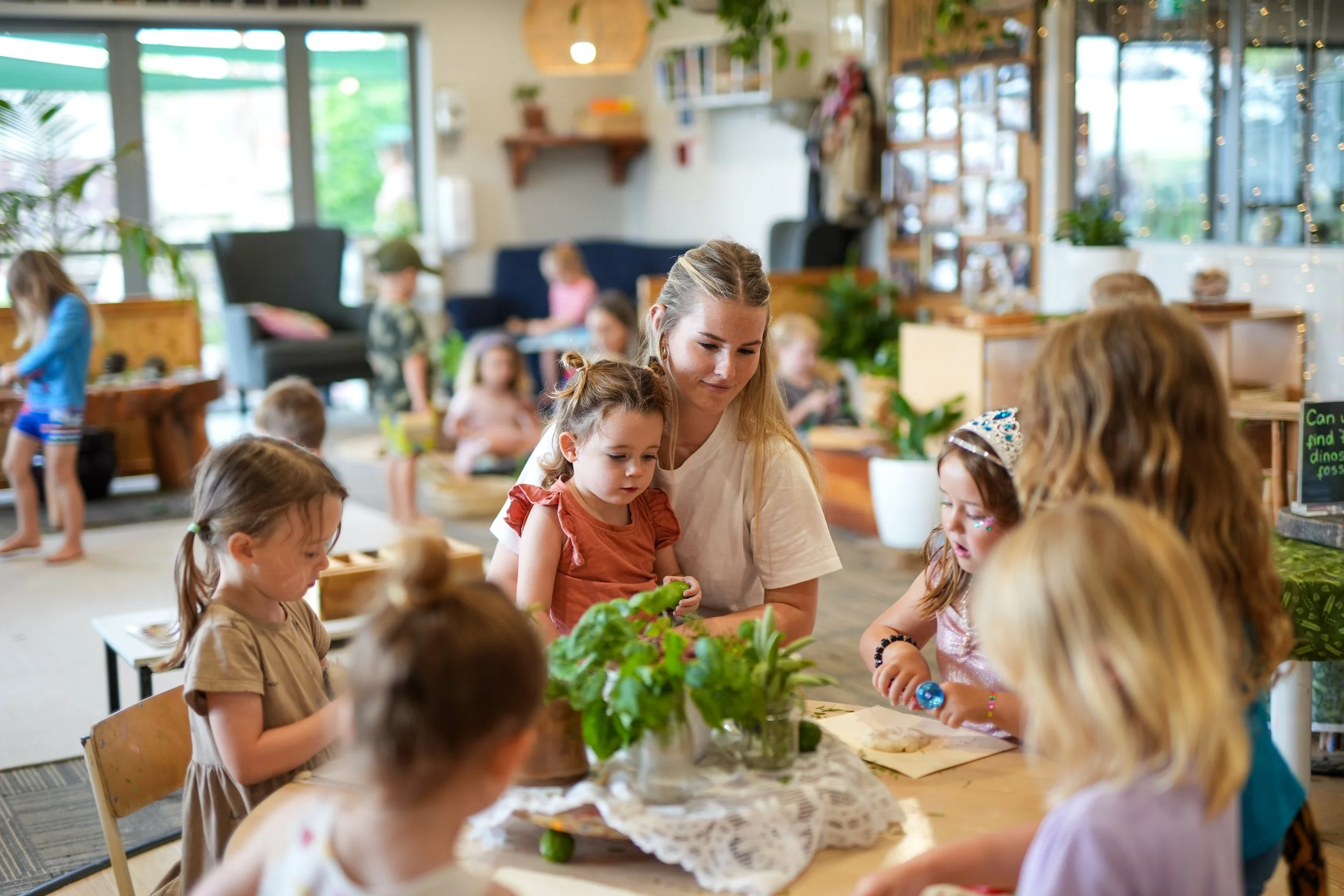 A group of young children and a woman are gathered around a table in a bright, cozy indoor space. The children are engaged in an activity, with some wearing princess crowns and colorful accessories. The atmosphere suggests a children's party or educa