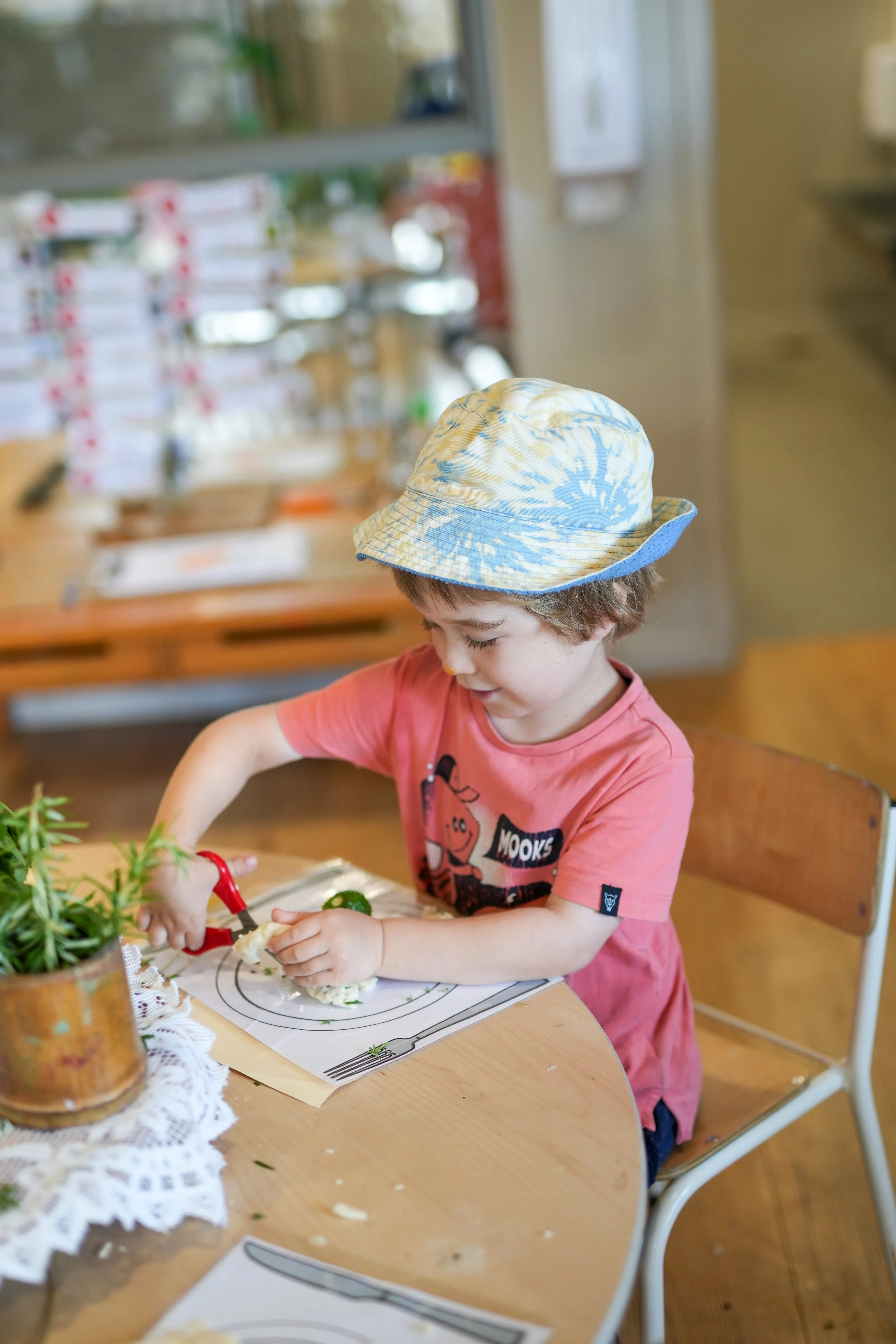 A young boy wearing a tie-dye bucket hat and a pink t-shirt is sitting at a wooden table, using a toy knife to cut food on a plate, with a potted plant and a lace doily nearby.