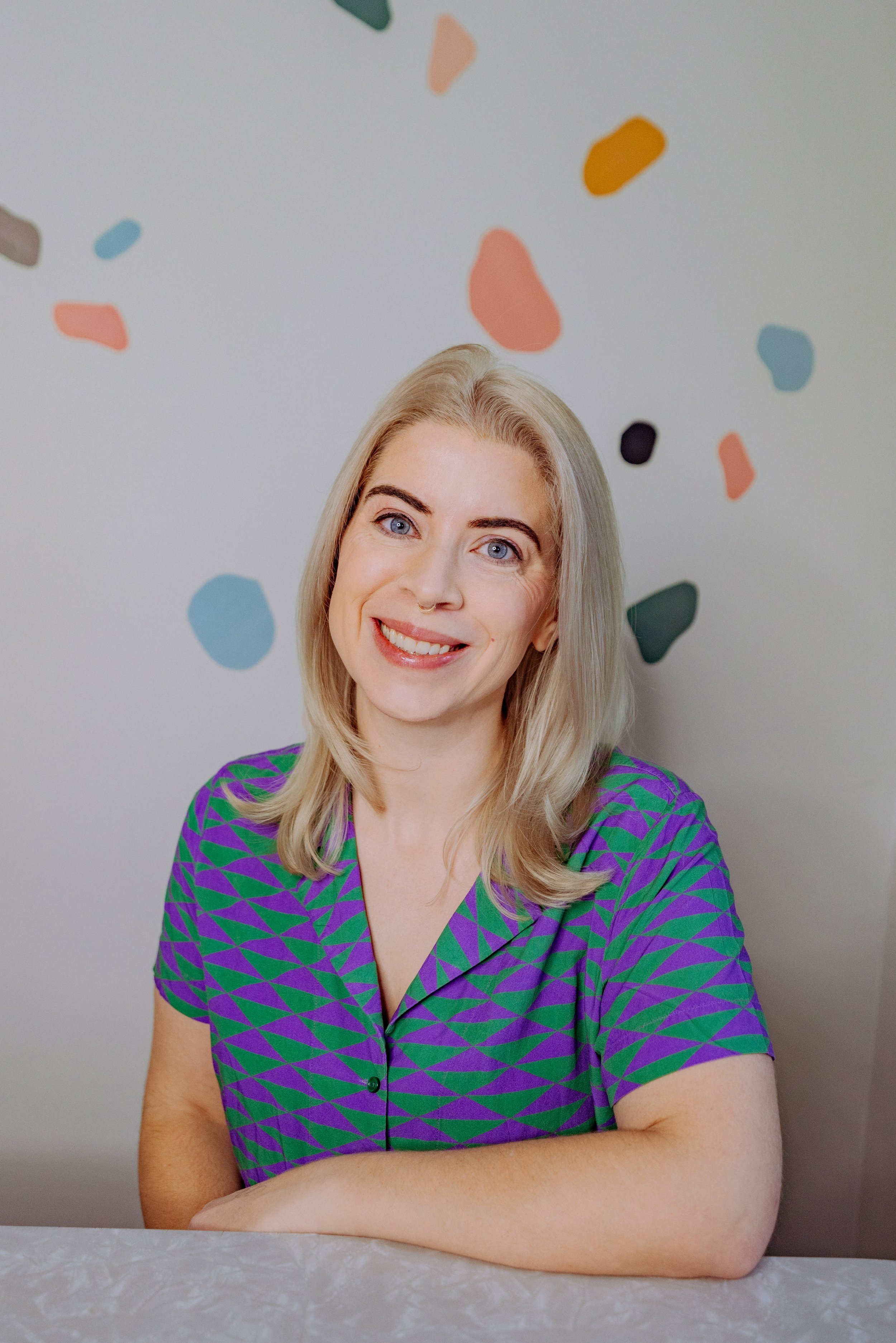 A smiling woman with blonde hair and blue eyes, wearing a purple and green geometric patterned shirt, sitting at a table with colorful abstract shapes on the wall behind her.