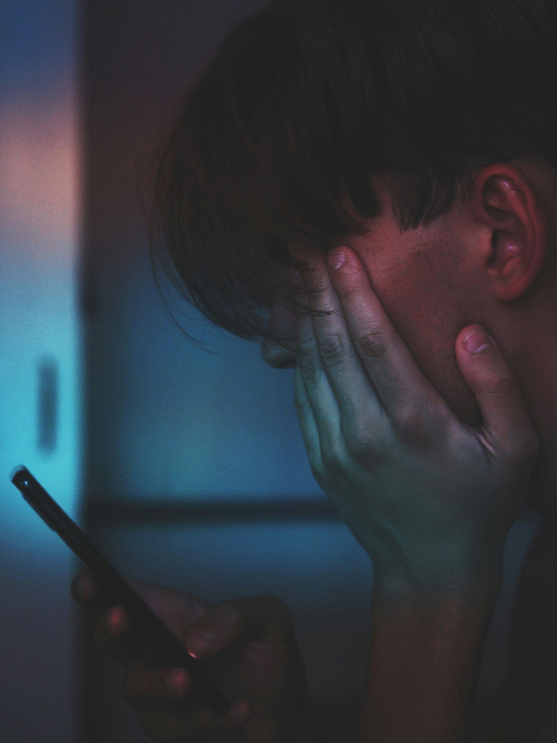 A person with dark hair and skin holds their face with one hand while looking at a smartphone in a dimly lit environment, appearing distressed or worried.