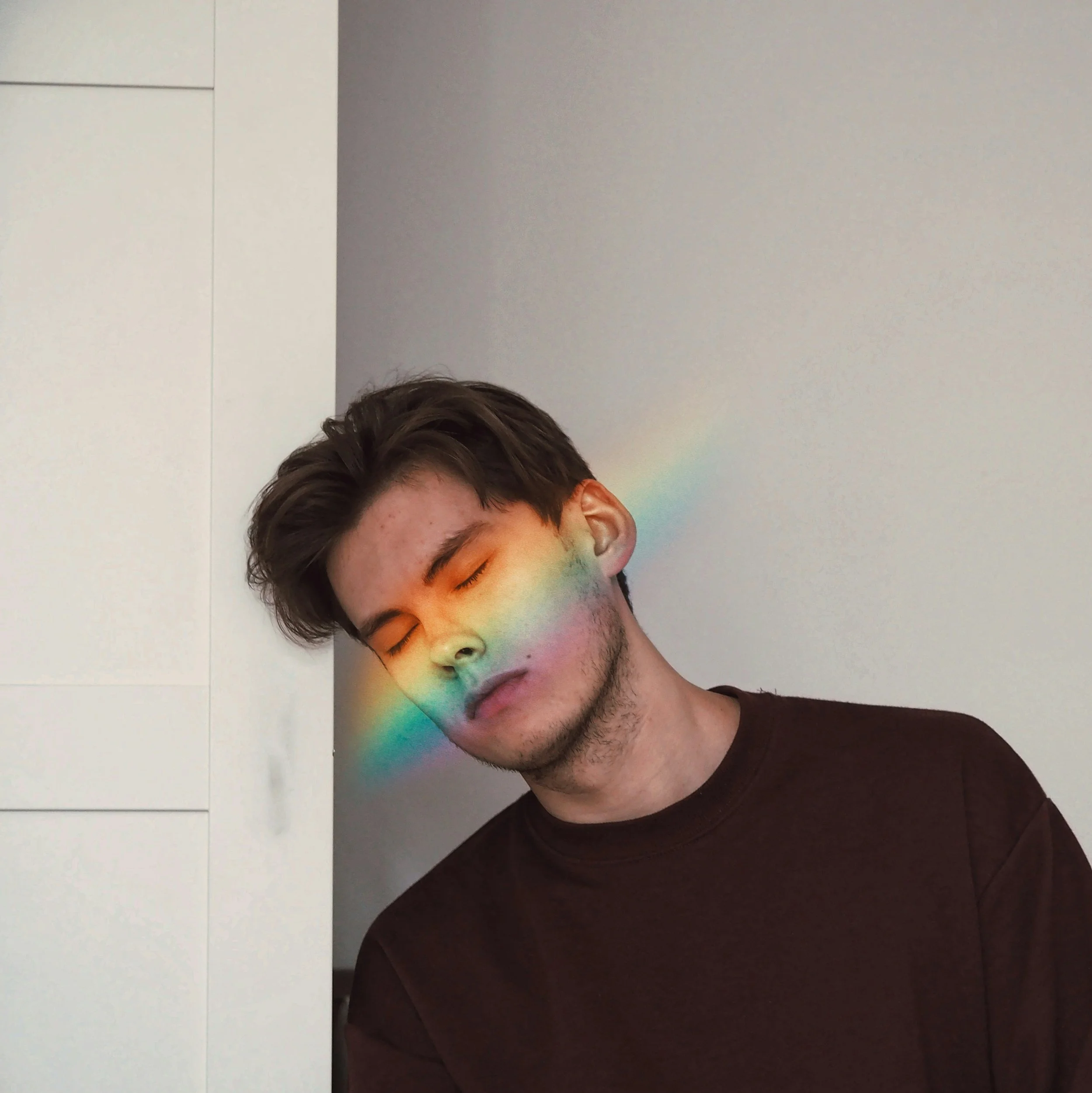 Young man with dark hair and stubble, eyes closed, leaning against a wall with a rainbow light streak across his face.