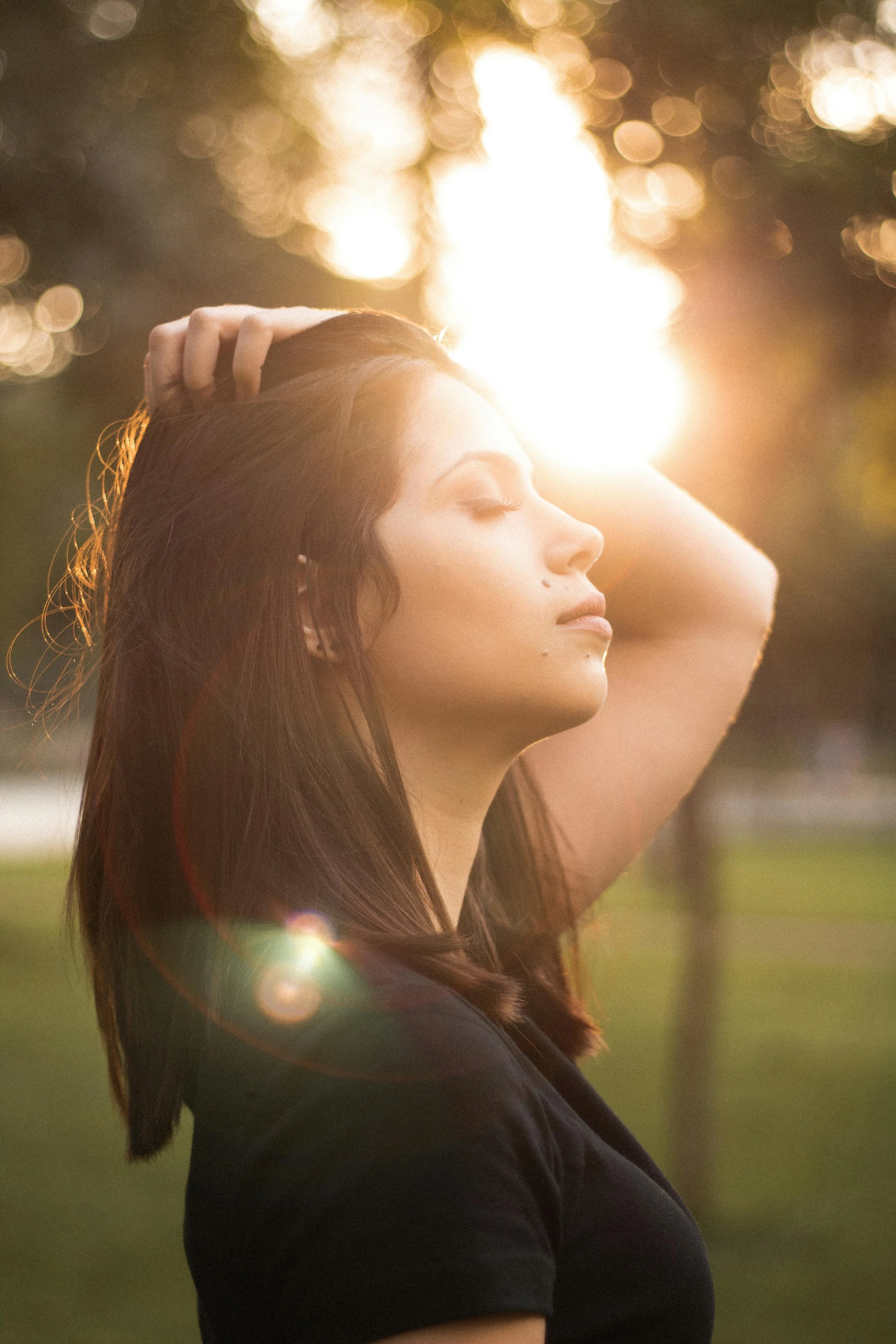 A woman with long dark hair, eyes closed, and one hand on her head, stands outdoors during sunset with a bright sun behind her.