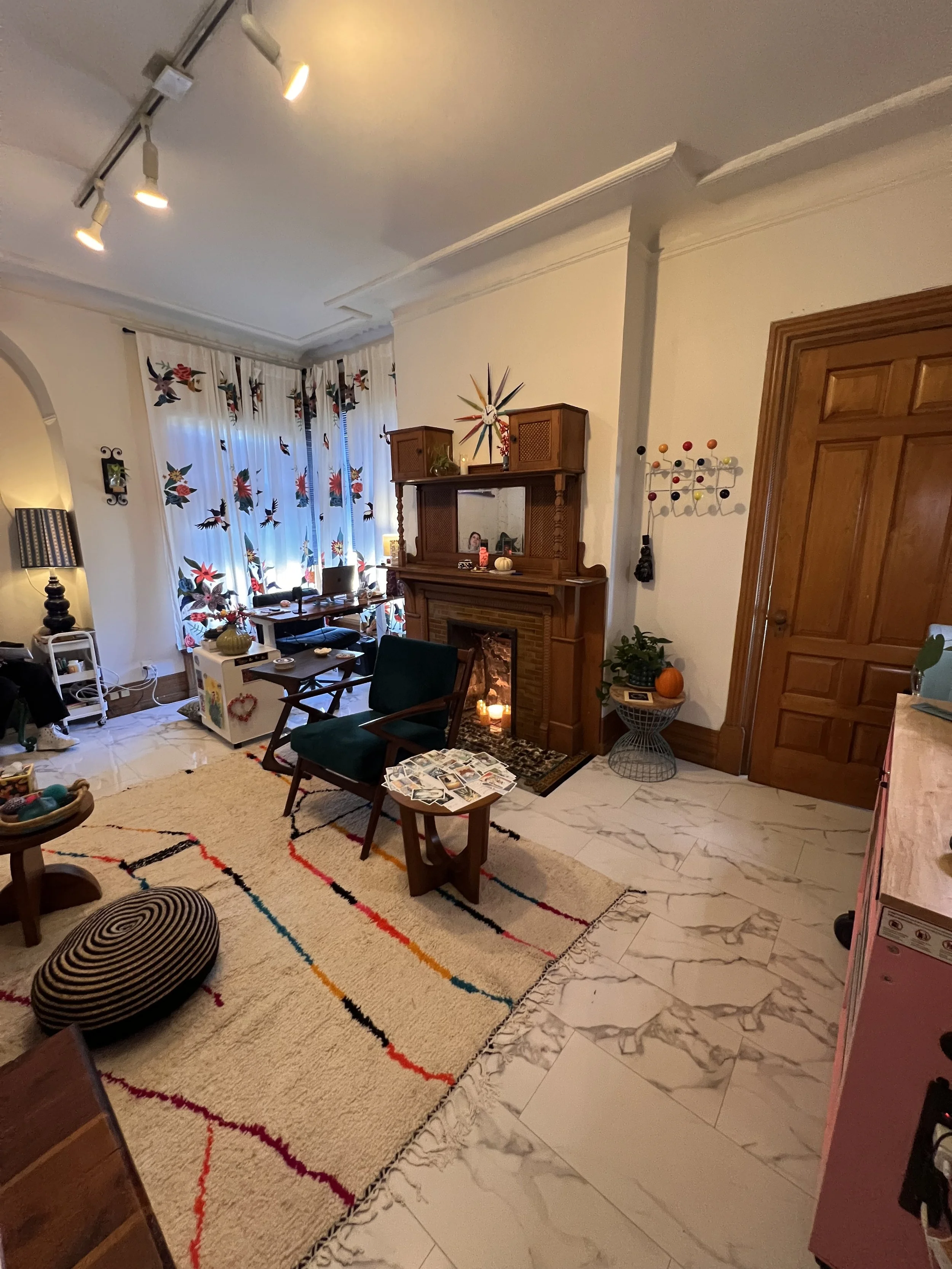 A cozy living room with a colorful striped rug, a fireplace, and a window with decorative curtains featuring flowers and birds.