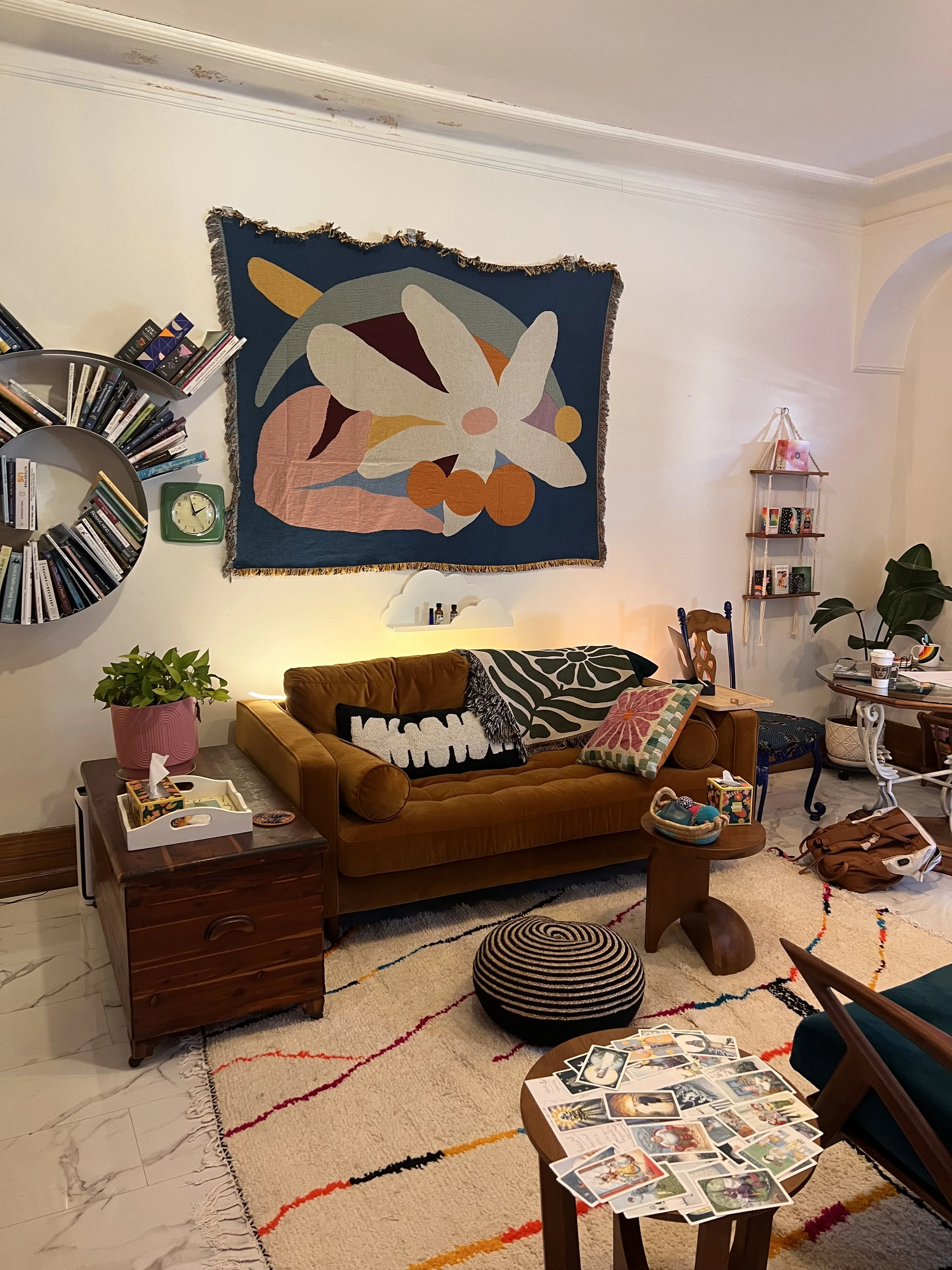 Living room with a mustard-colored sofa, a patterned pillow, a small side table with a tray of objects, a rug with colorful lines, a black and white striped pouf, and a wall tapestry with an abstract floral design. There are bookshelves, plants, and various furniture and decorative items.