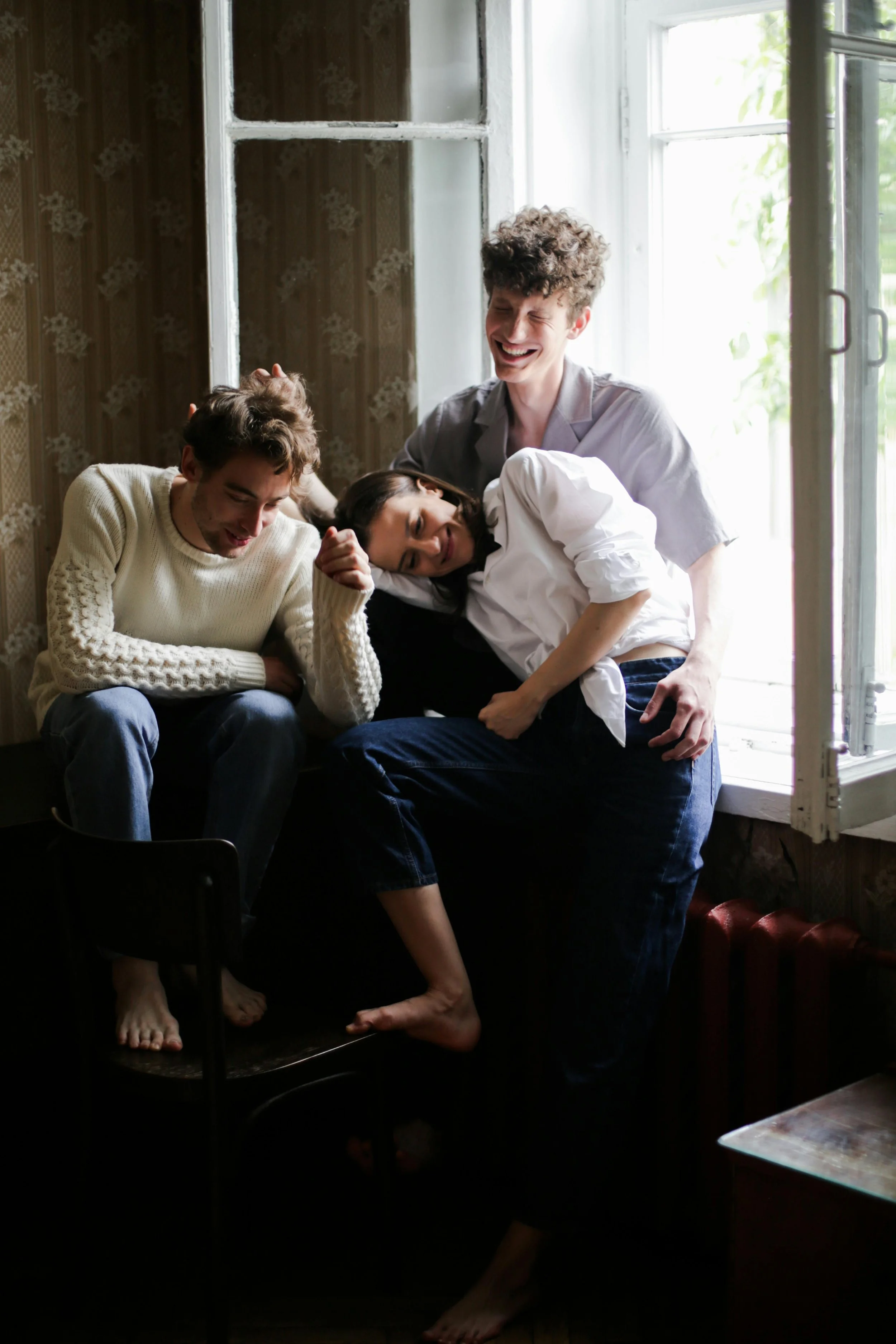 Four young adults sharing a joyful moment by a window with sunlight, sitting on a window sill, with one leaning on another and smiling.