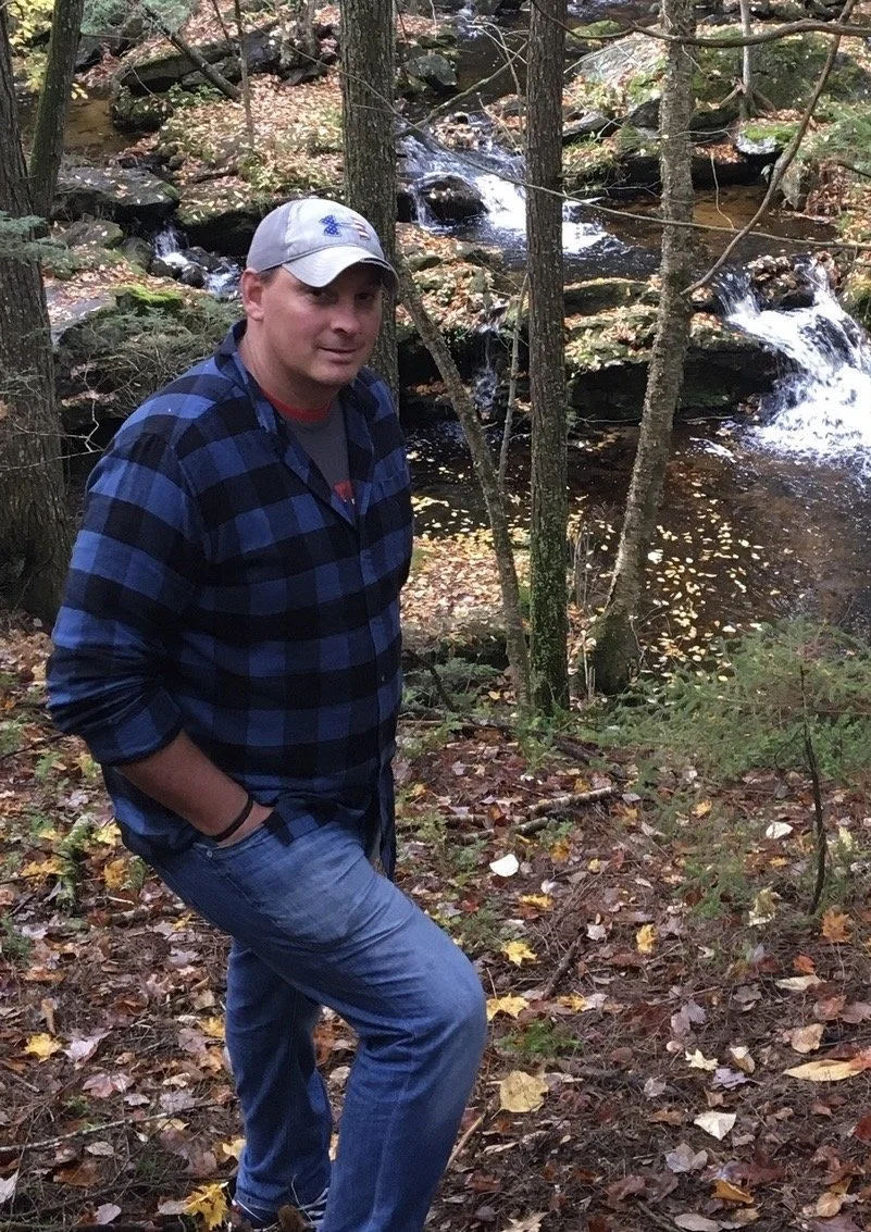 A man wearing a gray baseball cap, blue and black plaid shirt, and jeans stands in a forested area with fallen leaves, near a small stream with rocks and flowing water.