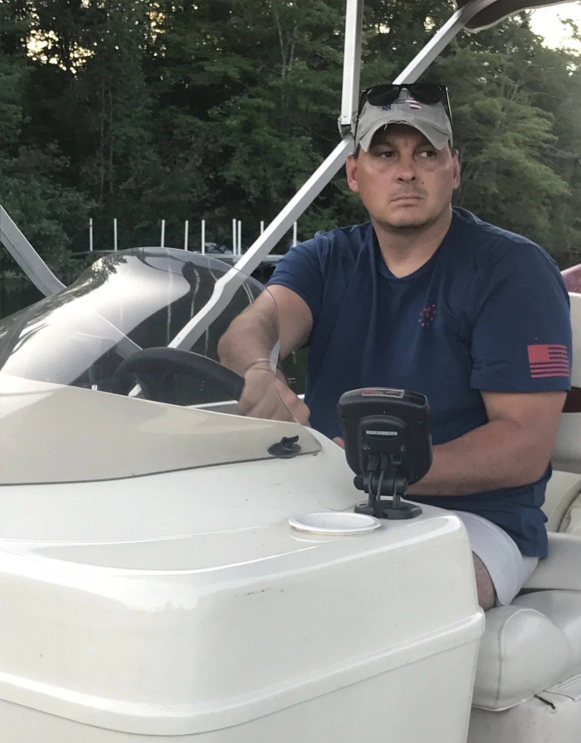 A man sitting in a white boat wearing a navy blue t-shirt, a gray cap with sunglasses on it, and appearing serious, holding a device. There is a GPS or fish finder on the boat near him and a background of green trees.