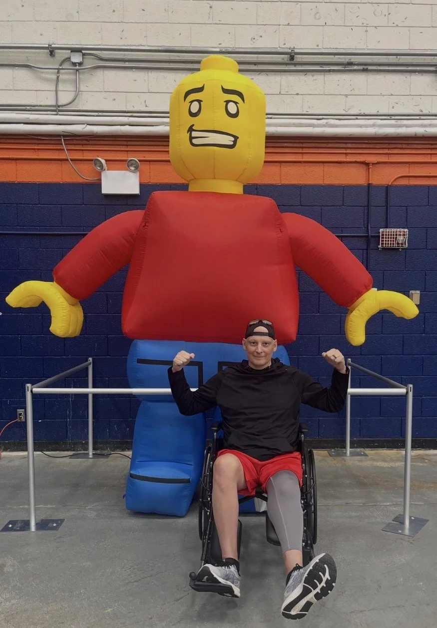 A man in a wheelchair flexing his biceps in front of a large LEGO figure with a yellow head, red shirt, and blue pants, inside a recreational or gym facility.