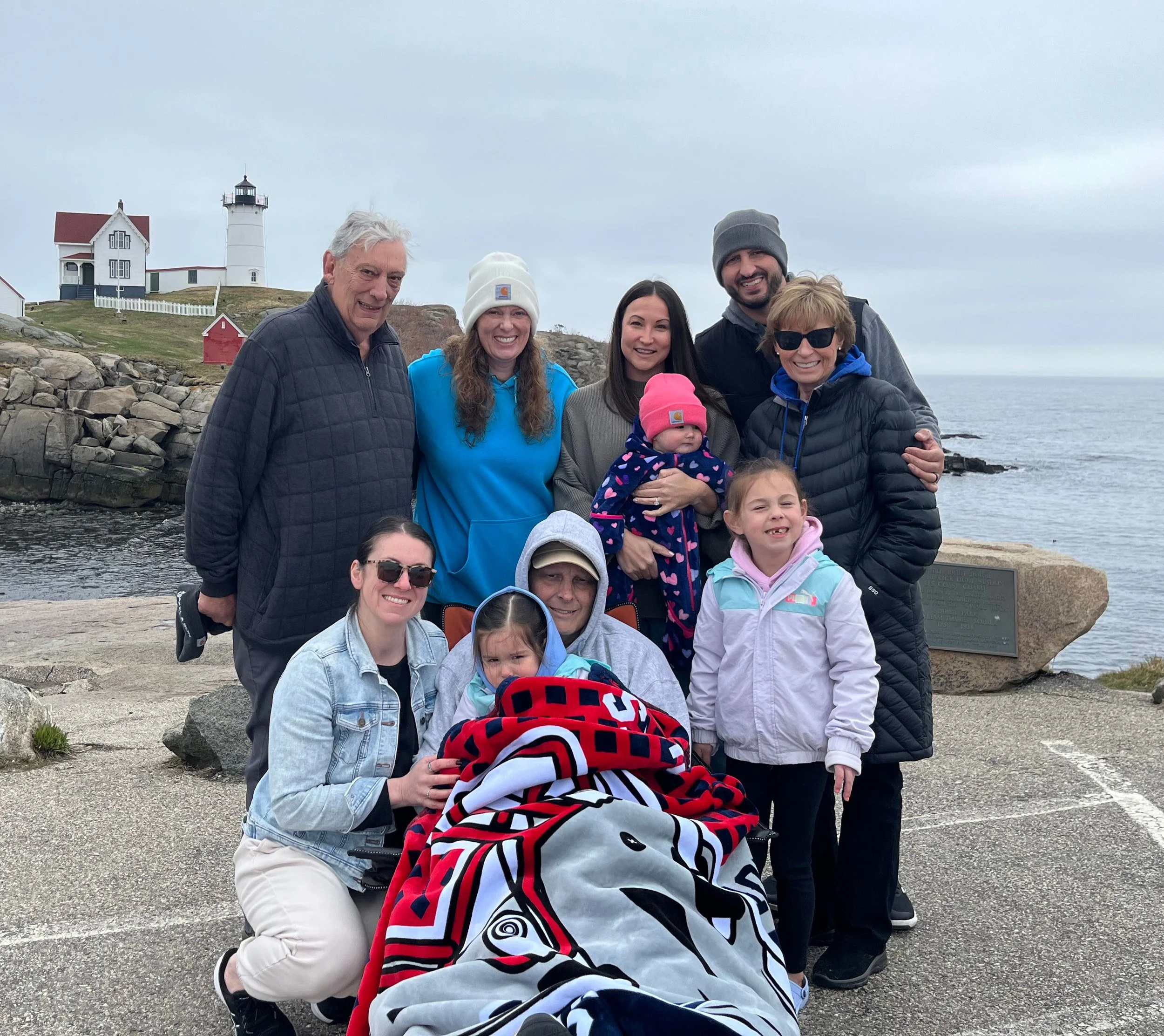 A group of nine people, including children, posing outdoors near a rocky shoreline with a lighthouse and lighthouse keeper's house in the background during overcast weather.