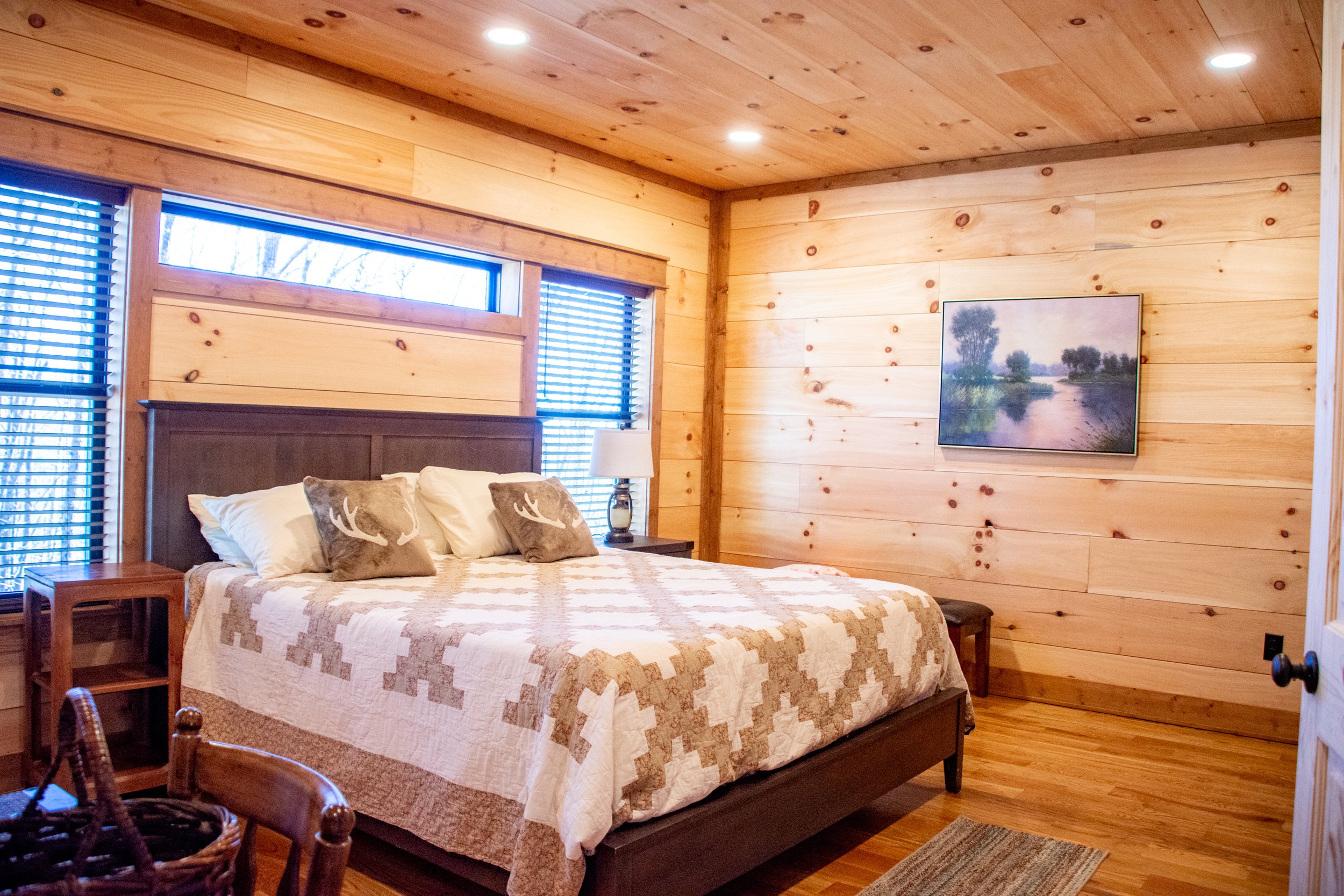 A cozy bedroom with wooden paneled walls and ceiling, a large bed with white and beige bedding, decorative pillows featuring deer antlers, a side table with a lamp, and a landscape painting on the wall.