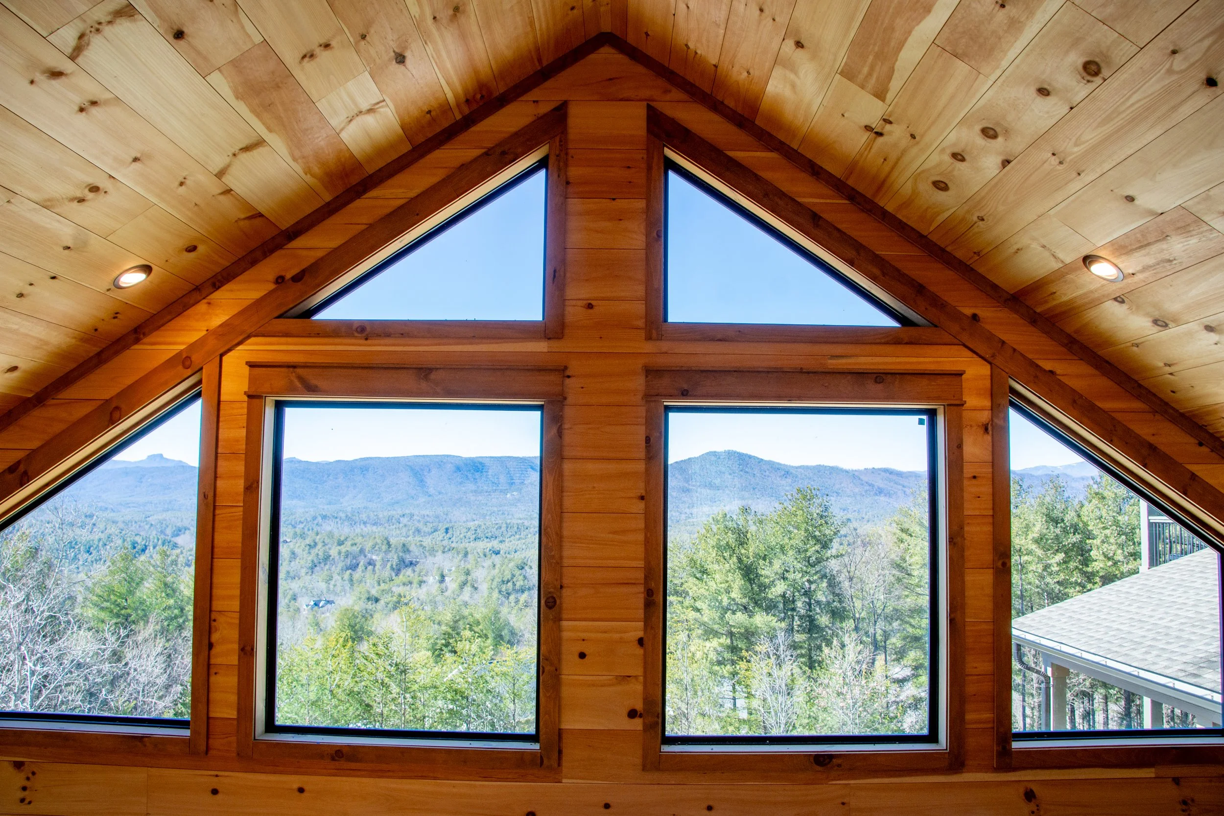 Interior view of a wooden house's ceiling and large windows showing a forested mountain landscape outside.