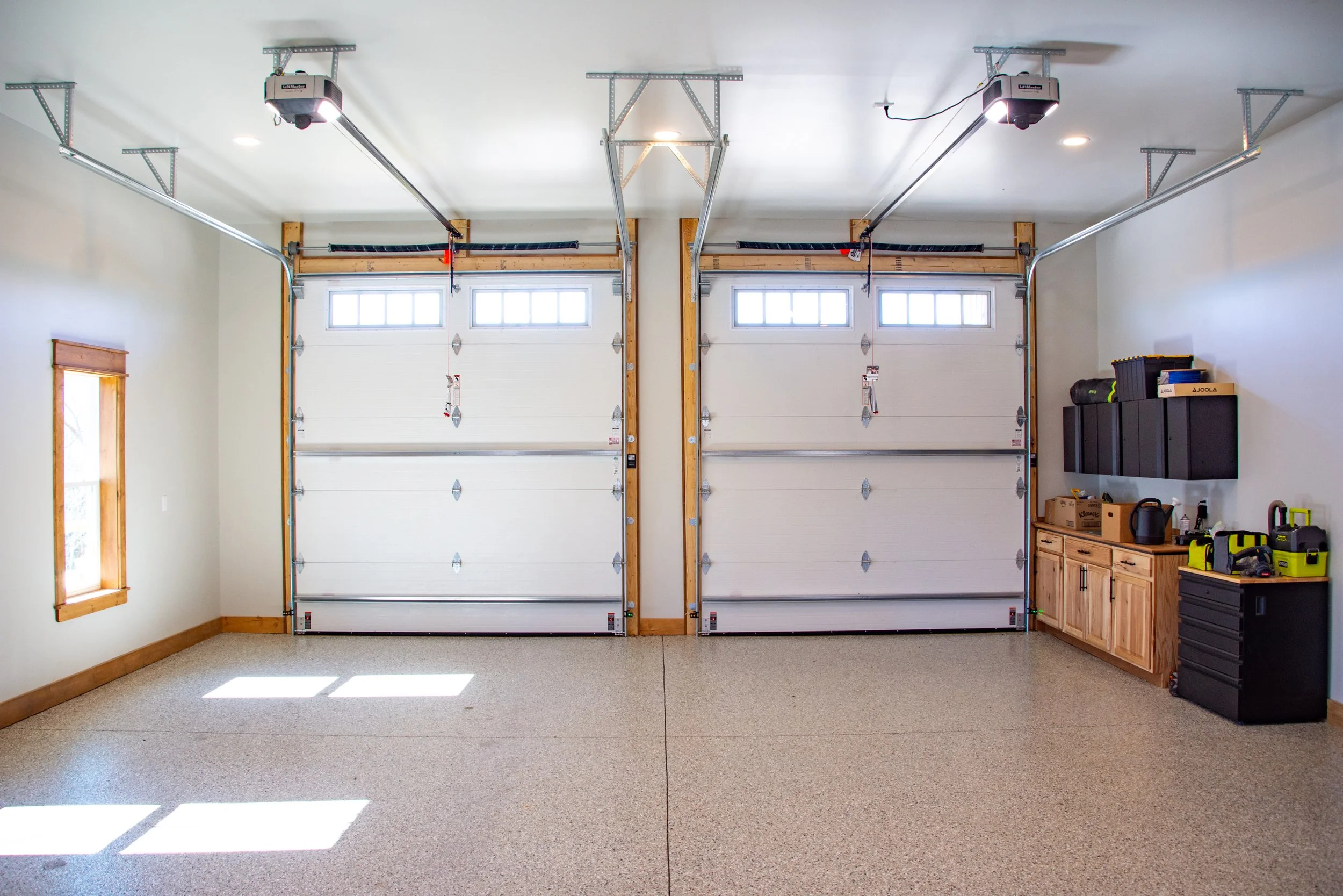A clean two-car garage with two white garage doors, a window on the left wall, and a tool storage area on the right with cabinets and various tools.