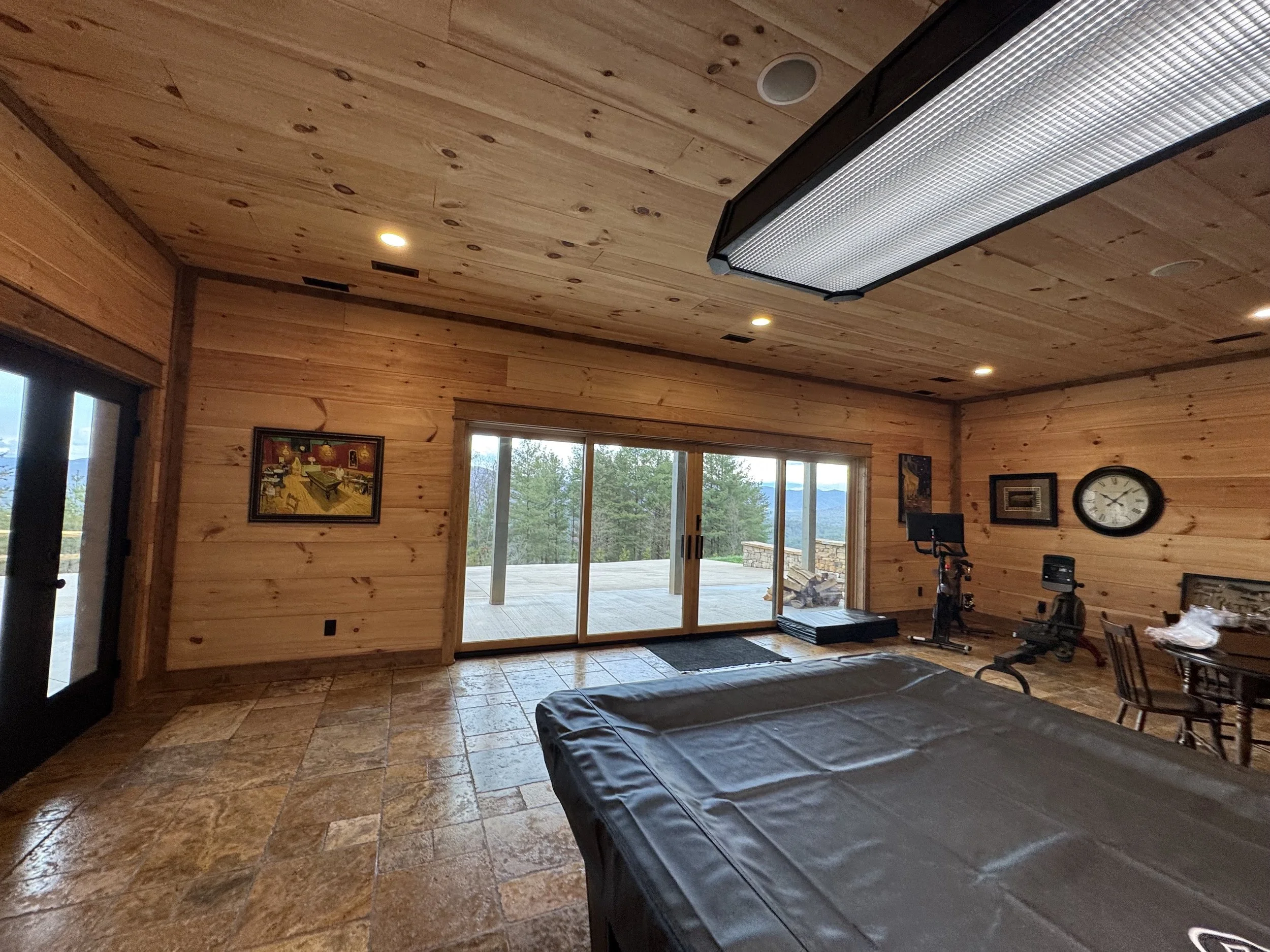 A spacious room with wooden walls and ceiling, featuring a large sliding glass door leading to an outdoor deck with a mountain view, and an interior with exercise equipment, a clock, and artwork.